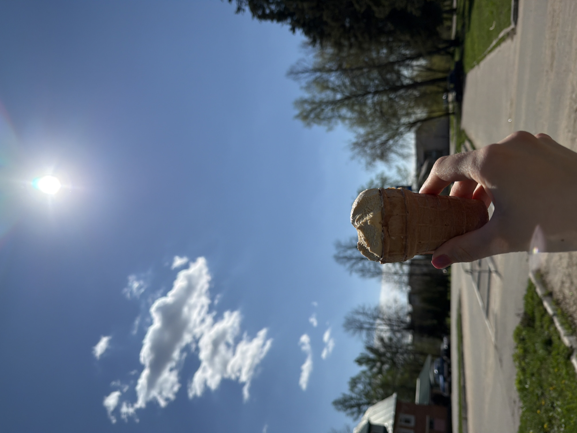 A hand holds a partially eaten ice cream cone against a backdrop of a sunny blue sky with scattered clouds. The foreground includes the ice cream cone and the holding hand, and beyond, a street scene with trees, and building rooftops are visible. Based on the location provided, this scene could be a street view in Reshetnikovo, Russia, captured on a sunny day.