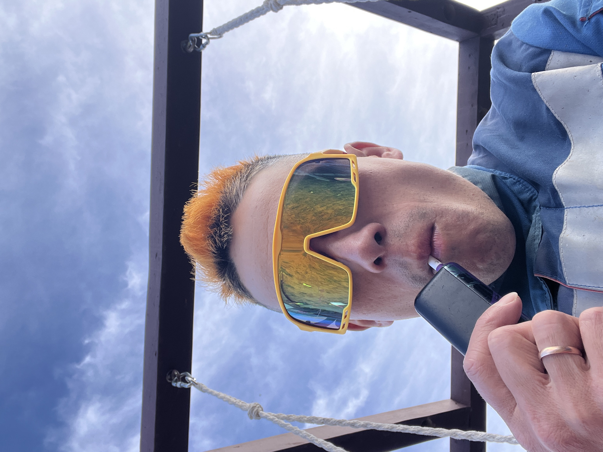 A person is viewed from a low angle, holding a dark-colored electronic device to their mouth. They wear bright yellow sunglasses and have orange-dyed hair. The person is wearing a blue jacket with white accents and a wedding ring on their hand. They are standing beneath a brown wooden structure with ropes and metal hardware. The sky is visible in the background. The image does not explicitly state the location is in Chaikovsky, Russia.