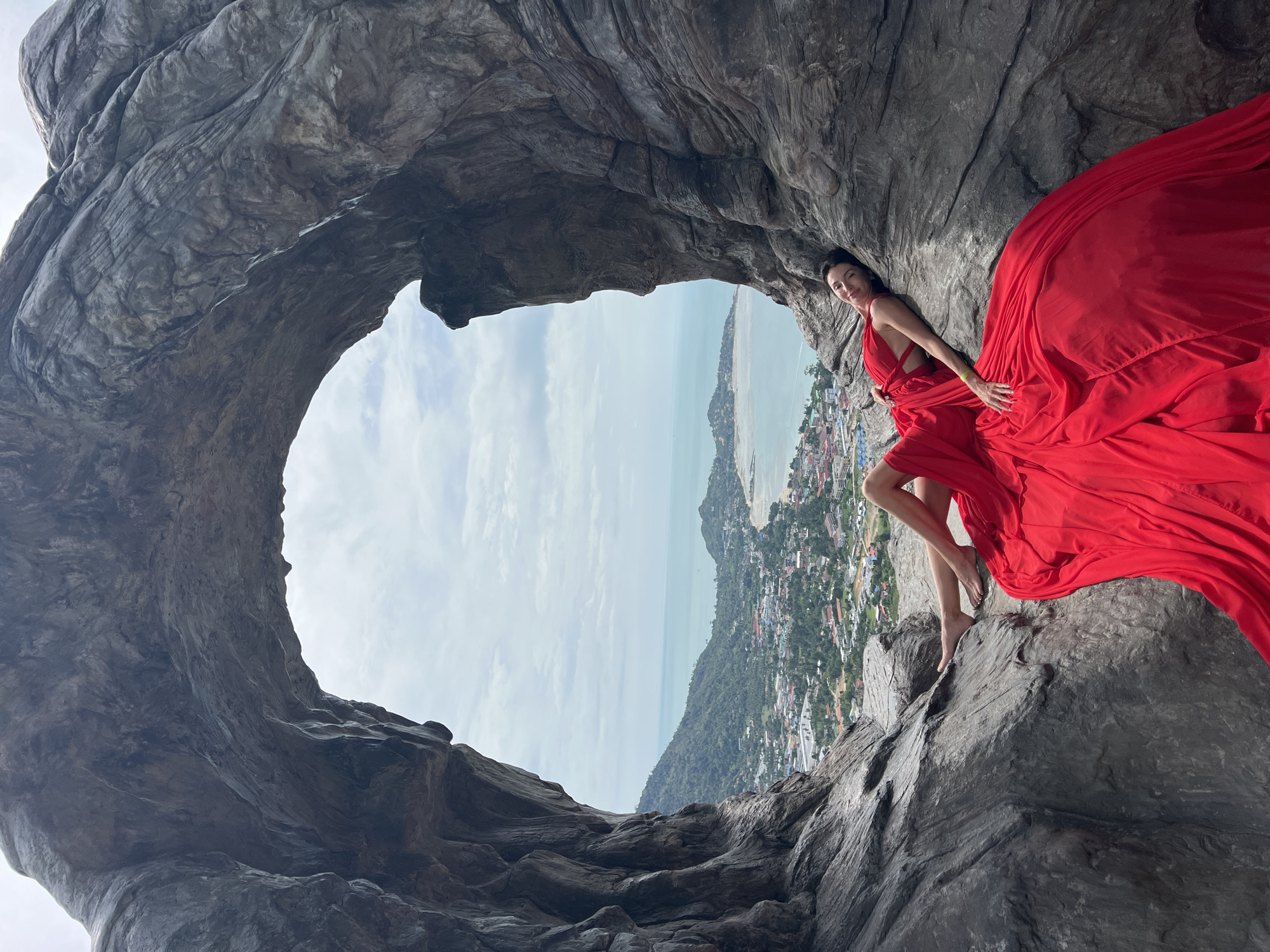 The image is taken at Overlap Stone, Baan Hua Thanon, Thailand. It shows a woman in a red dress lying on the rocks. She is inside a large stone hole with a view of the coastline and city in the background. The sky is partly cloudy.