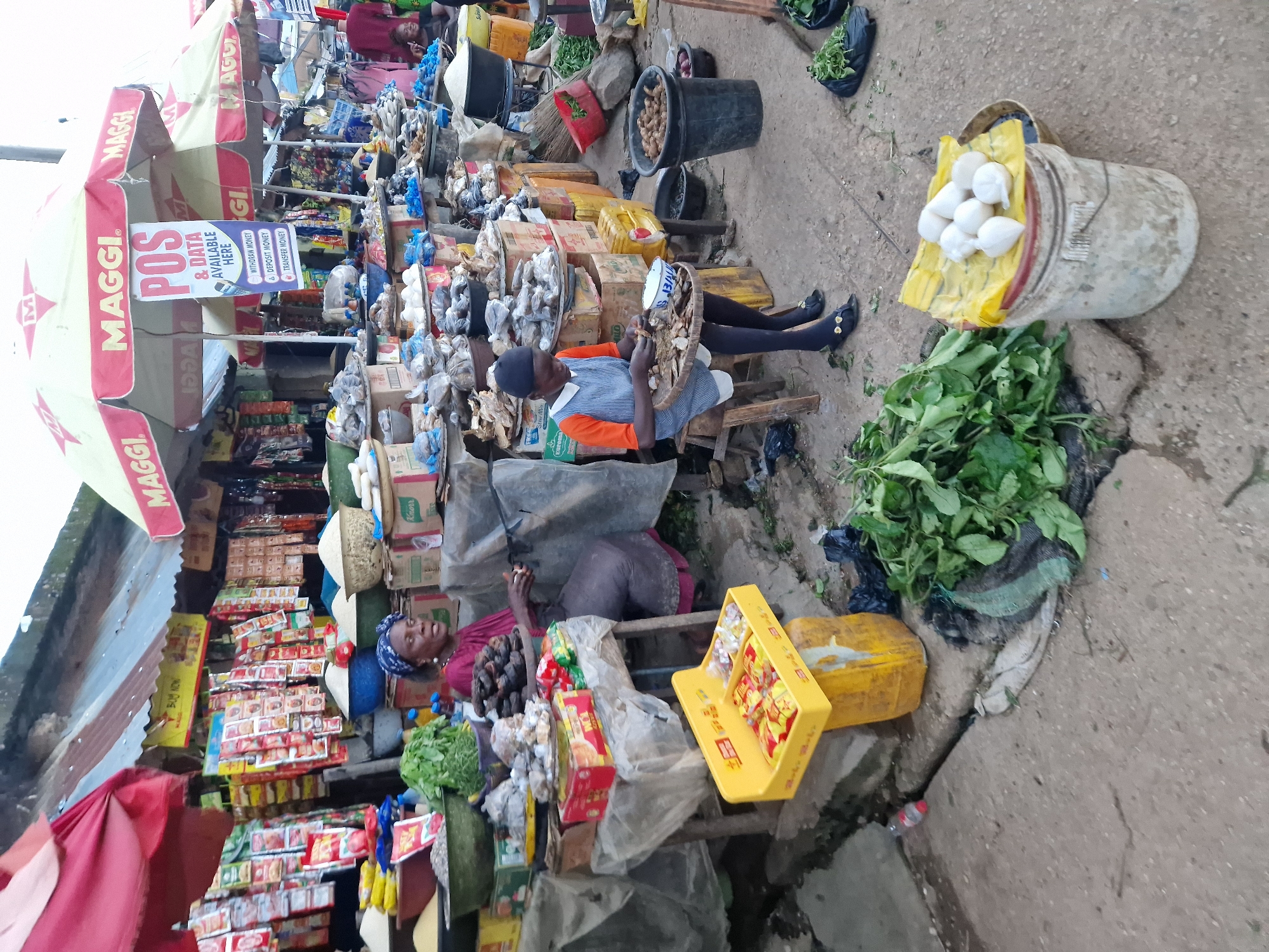 An outdoor market scene in Kabong, Nigeria, depicts a busy commerce area with vendors and various goods. Two individuals, likely vendors, are visible. The first, an adult woman wearing a purple dress and a blue headwrap, sits behind a table laden with produce and packaged goods. Her gaze is directed towards the right, and her hands appear to be engaged in some activity. To her left, a younger person, dressed in an orange shirt and a blue and white striped apron, sits on a stool. This individual is actively sorting through dried fish displayed on a round wooden tray.
The market stall features an array of products including rice, vegetables, packaged food items such as Maggi seasoning, and what appear to be dried fish and yams. Several umbrellas, branded with "MAGGI," provide shade over the stalls. A sign advertising "POS & DATA AVAILABLE HERE" is prominently displayed on one of the umbrellas, detailing services like withdrawal, deposit, and transfer of money. The ground is dirt and concrete, with some scattered debris, indicating a typical bustling marketplace environment. In the foreground, a bucket topped with a yellow cloth holds several white, egg-shaped objects, possibly eggs, and a pile of fresh green leafy vegetables lies beside it.