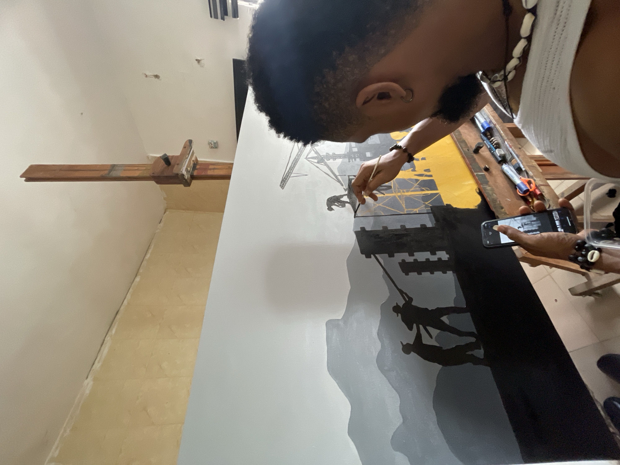 A person with dark hair and skin is shown painting on a large canvas. The painting depicts silhouetted figures on a craggy landscape. Above the landscape, there are abstract structures resembling scaffolding or industrial cranes. A second person stands next to the artist and is taking a photo of the artwork with a smartphone. Tubes and other art supplies are placed on a surface near the canvas. A wooden easel is visible in the background. The location is in Ado, Nigeria.