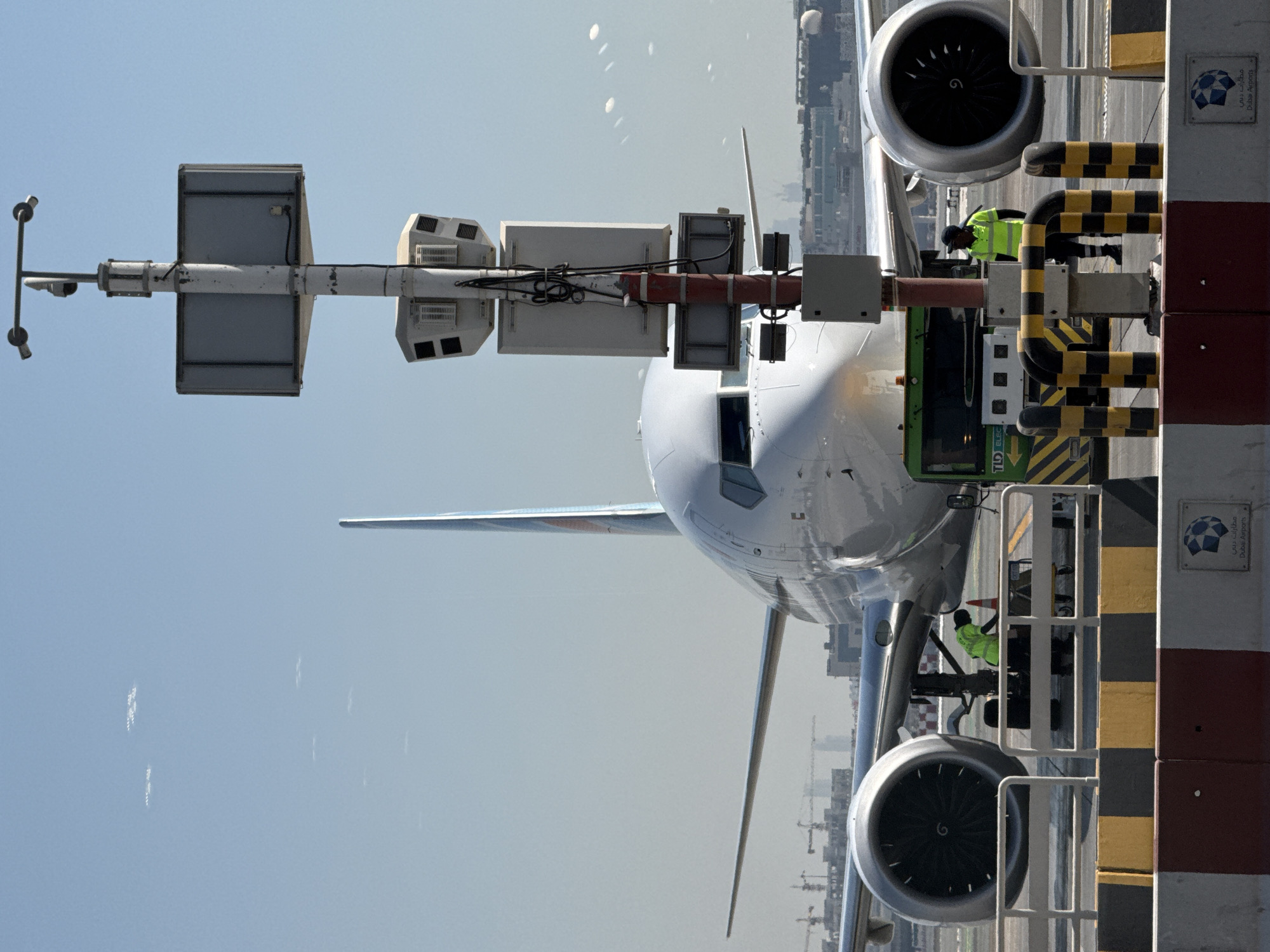 An outdoor, ground-level, wide shot shows an airplane on an airport tarmac in Dubai, United Arab Emirates, under a bright, clear blue sky. The airplane, a passenger jet with white fuselage and two engines visible, is the primary subject. The nose of the plane is centered in the frame, with one of its engines prominent on the left. A tall, gray pole with multiple cameras and electronic devices is situated to the right of the plane. In the background, construction cranes and buildings are visible. In the foreground, there is a yellow and black striped barrier, and two individuals in high-visibility yellow vests are near the plane, one appearing to be working on it and the other standing beside a vehicle. A green tow truck is positioned in front of the plane's landing gear. The overall scene depicts airport operations on a sunny day.
