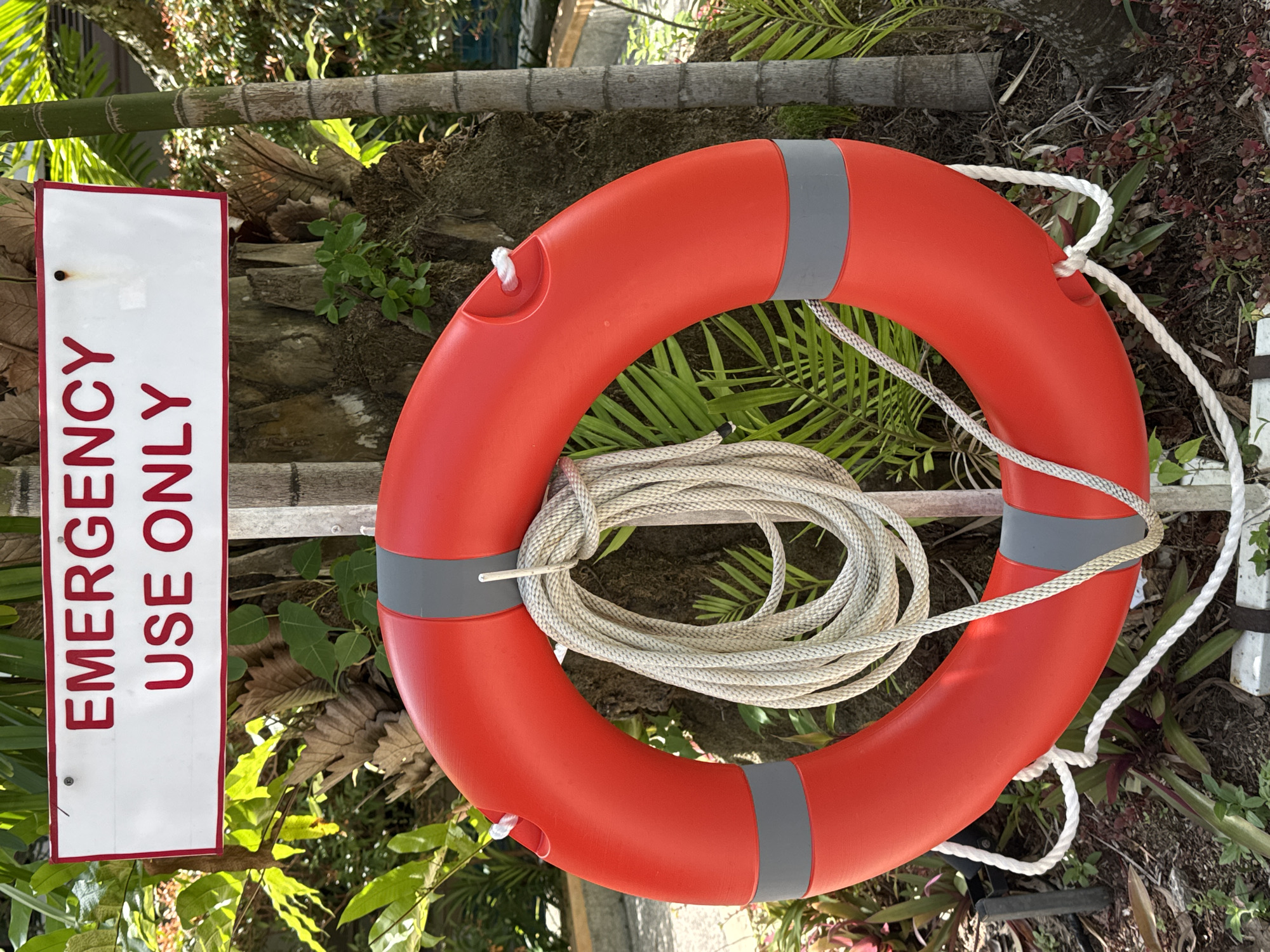 A bright red life preserver with a coiled white rope is hanging on a metal post in front of a palm tree. Above the life preserver, a white sign with red lettering reads "EMERGENCY USE ONLY." The background consists of lush green foliage, suggesting an outdoor location. The ground is covered in dirt, small plants, and some fallen leaves. The scene is brightly lit, indicating daytime. The location is identified as Karon, Thailand. There are no people or animals present in the image, nor are there any actions or interactions depicted.