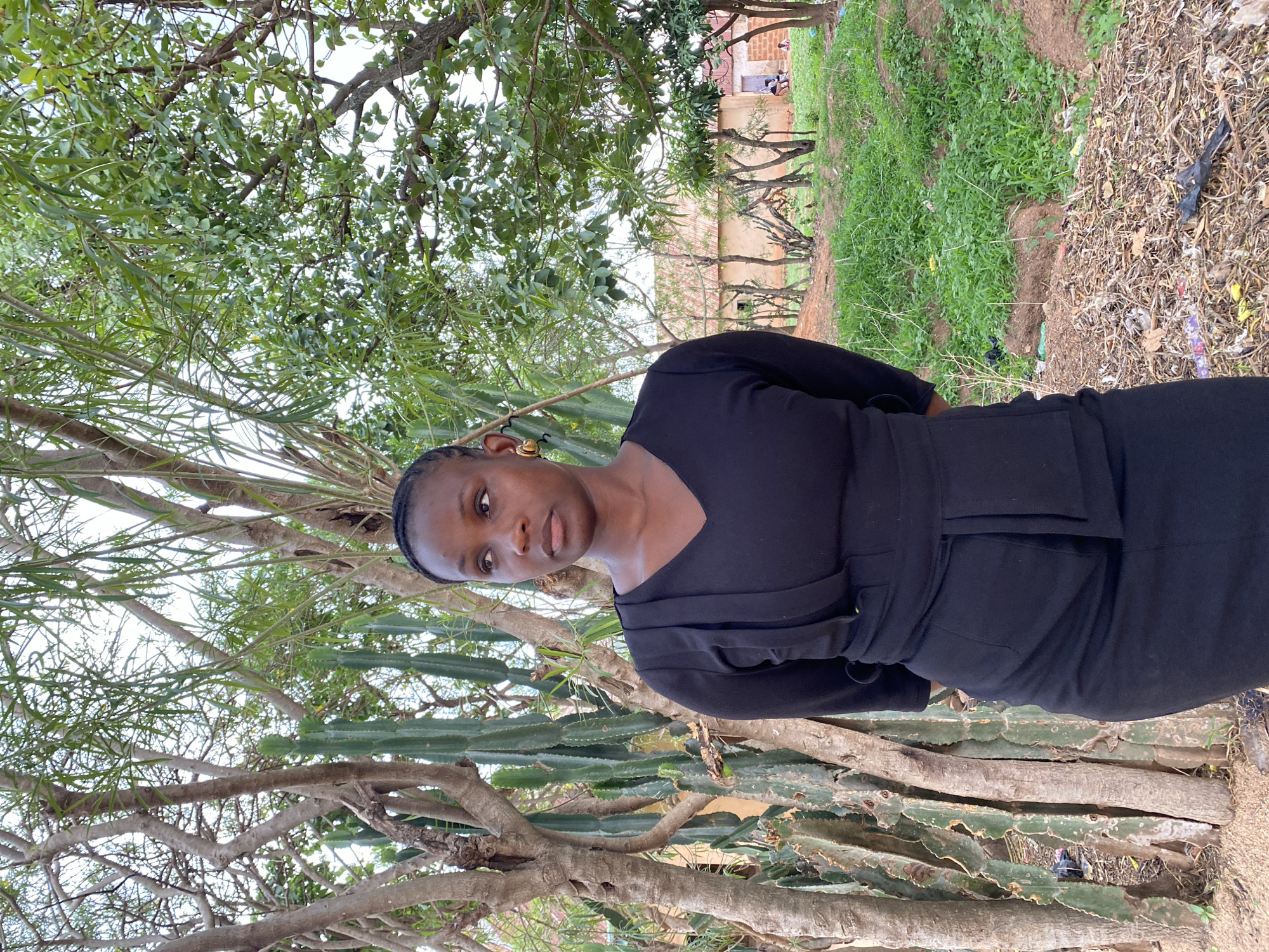 The image shows a woman in a black dress standing in an outdoor environment in Kuru Dungus, Nigeria. Behind her are trees with green foliage and a cactus-like plant. In the background, there's a building with visible individuals near a door. The ground consists of grass, vegetation, and scattered debris.