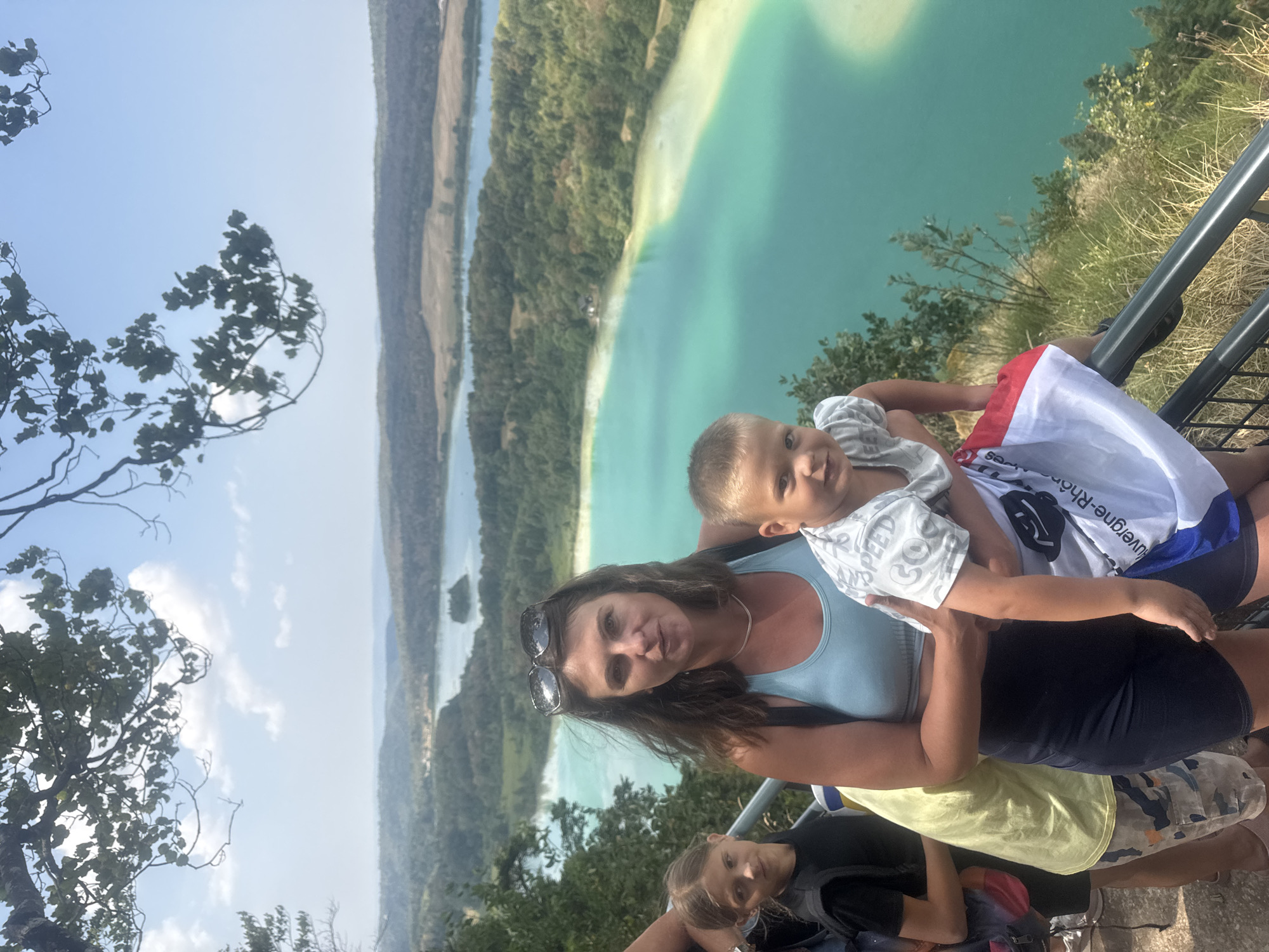 This media file captures a family enjoying a scenic overlook on a sunny day. The subjects are a woman and three children, two boys and a girl. The woman, positioned in the foreground, is wearing sunglasses on her head and a light blue tank top. She is holding one of the young boys, who is wearing a grey and white striped shirt. To her right, another child, a girl, is partially visible wearing a black shirt. Further down, another young boy is visible.
The group is standing near a metal railing, suggesting they are on a viewing platform or at a scenic lookout. Behind them, a breathtaking landscape unfolds. A large body of turquoise water, possibly a lake or reservoir, dominates the background, with lush green forests and hills surrounding it. The sky is clear and bright, indicating daytime with good weather. The image is oriented vertically, with the landscape appearing to stretch out above the people.
There is visible text on the white and blue flag held by one of the children, but it is partially obscured and difficult to read clearly. The overall mood appears to be joyful and relaxed, as the family takes in the beautiful view. The location context of La Chaux-du-Dombief, France, suggests this could be a popular tourist spot known for its natural beauty.