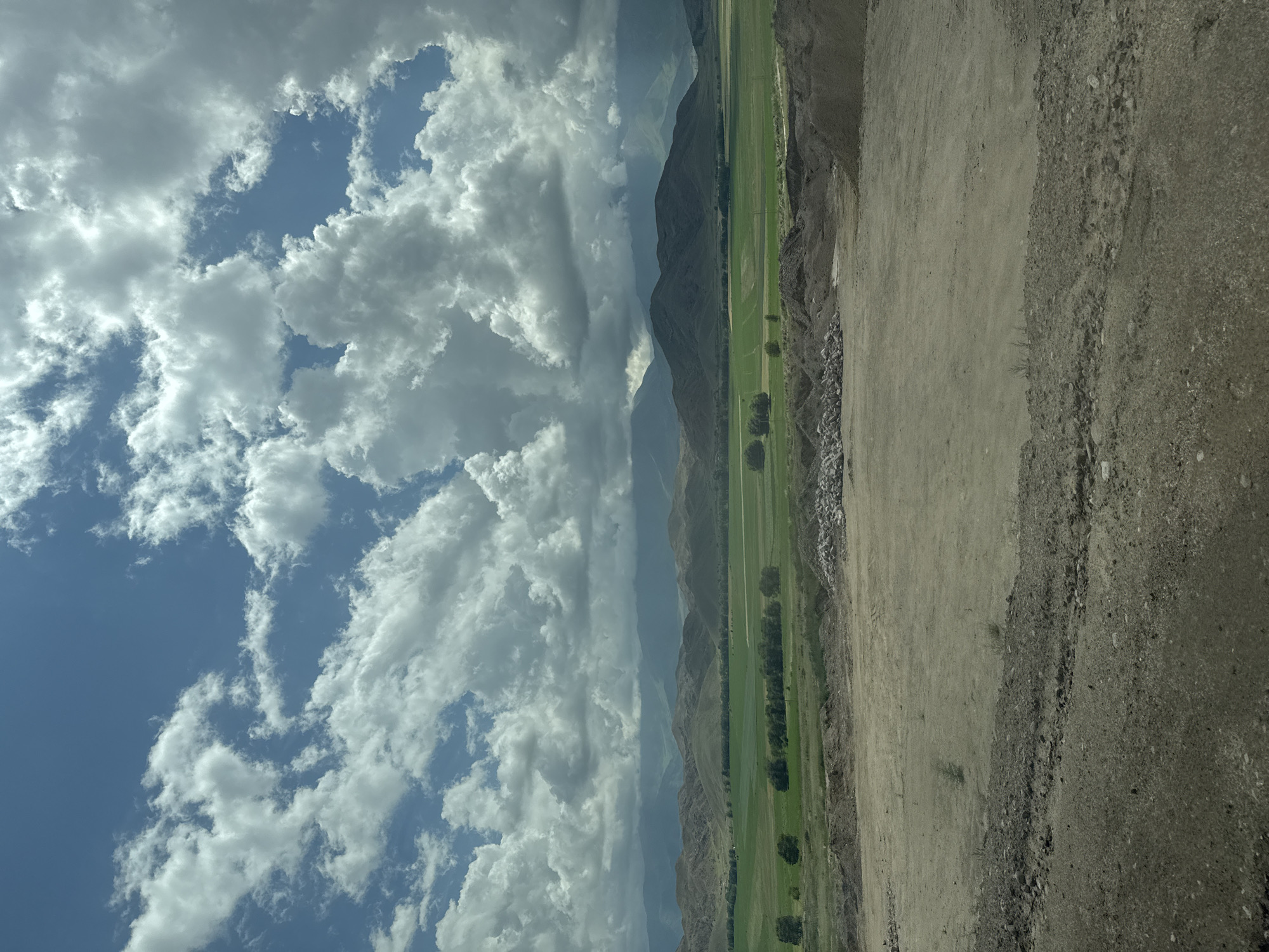 The image depicts an outdoor scene with a vast, partly cloudy blue sky dominating the upper portion. Below the sky, a range of rolling hills and mountains stretches across the horizon, with some appearing shadowed and others lighter in the distance. In the middle ground, a vibrant green field is visible, dotted with scattered trees and possibly some agricultural features. The foreground consists of a barren, dusty area with piles of dirt and rocks, suggesting a construction or excavation site. The overall environment appears to be rural or semi-rural, possibly on the outskirts of a settlement. The location is Ak Say, Kyrgyzstan.