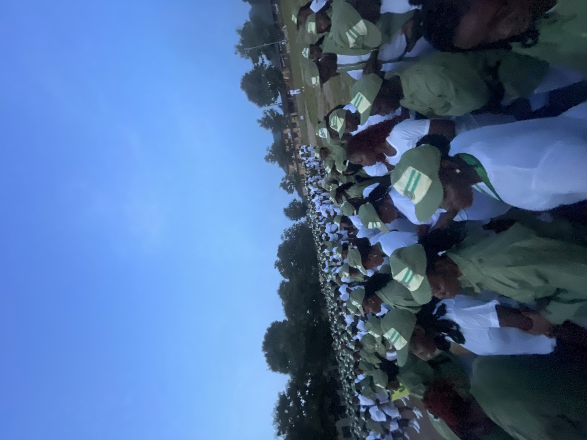 In the city of Ise - Ekiti, Nigeria, a large gathering of people, predominantly young adults, are assembled outdoors. They are dressed in matching light green uniforms with white accents and caps, suggesting an organized event or program. The individuals are standing close together, facing forward, and appear to be engaged in an activity, possibly a ceremony or a briefing. The background features a line of trees and buildings, indicating a semi-rural or institutional setting. The sky is a clear blue, suggesting it is daytime. The composition of the image shows a wide shot, emphasizing the scale of the group.