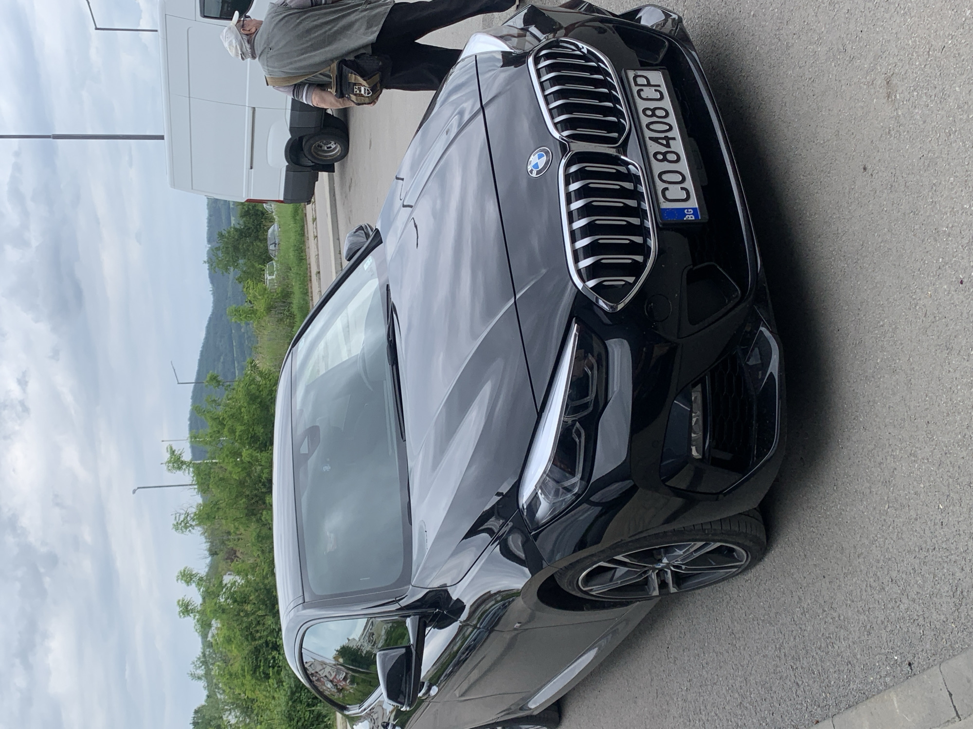 A black BMW 2 Series coupe is parked on a paved surface. Its front end is visible, showcasing its grille and headlights. The license plate reads "CO 8408 CP" with the "BG" insignia, indicating it is from Bulgaria, specifically the region of Sofia (CO). An individual wearing a light-colored cap, dark trousers, and a gray jacket is seen near the vehicle. A white van is partially visible in the background. The location appears to be near a road lined with greenery and streetlights, possibly in an urban area near Gabrovo, Bulgaria.