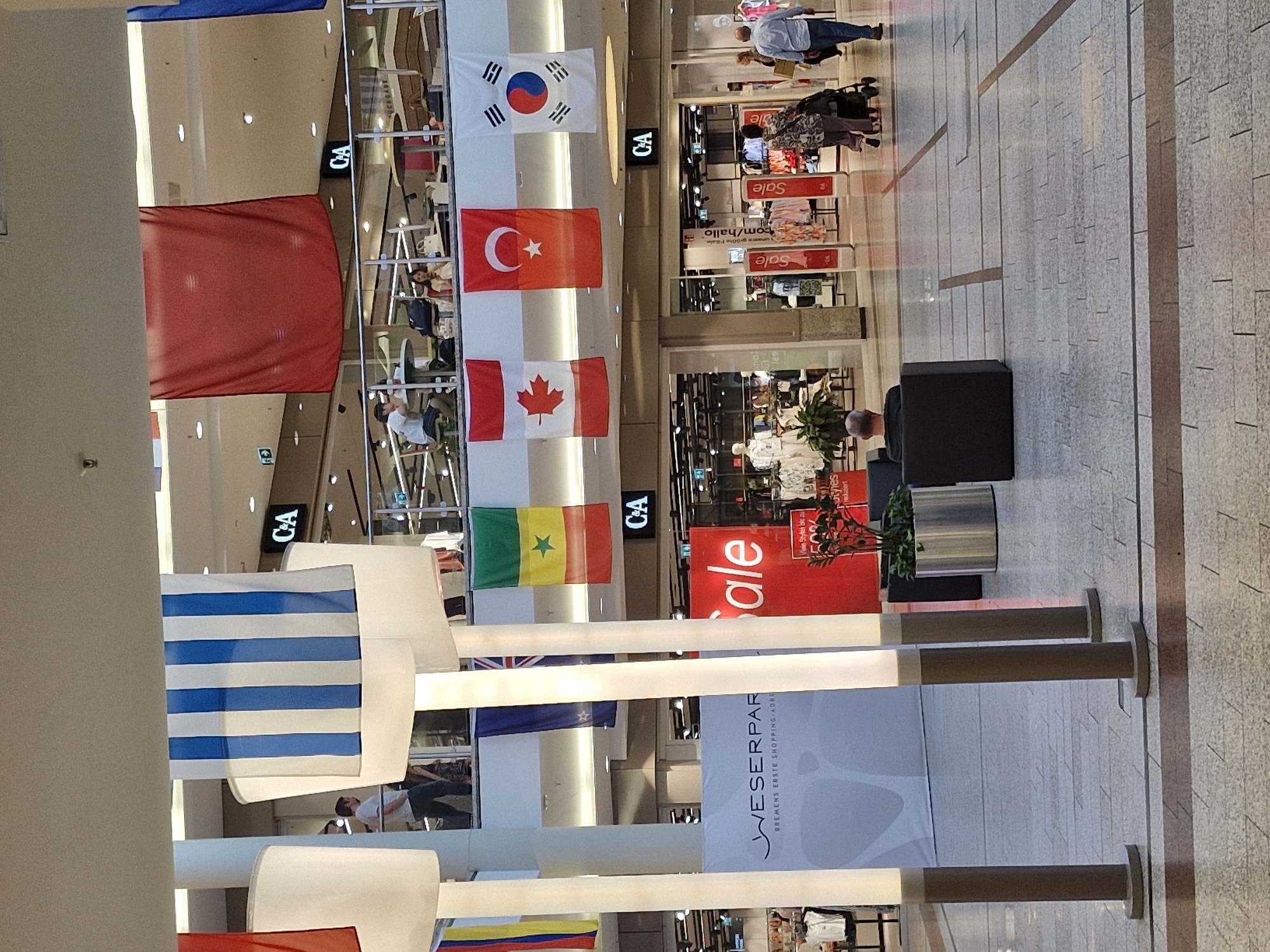 The image captures an interior view of the Weserpark shopping mall in Bremen, Germany. The scene depicts a wide, open-plan mall area with multiple levels visible. Large white light fixtures are positioned prominently in the foreground. Several national flags are hung from the ceiling, including flags of Israel, Canada, Turkey, South Korea, and Senegal, among others. Various shops are visible on the upper levels, with prominent signage for "C&A". In the lower level, a large red "Sale" banner is displayed. People are seen walking through the mall, and some are seated at tables on an upper level cafe area. One individual is seated in a dark cube-shaped chair in the foreground. The mall floor is tiled with a grey and brown pattern.