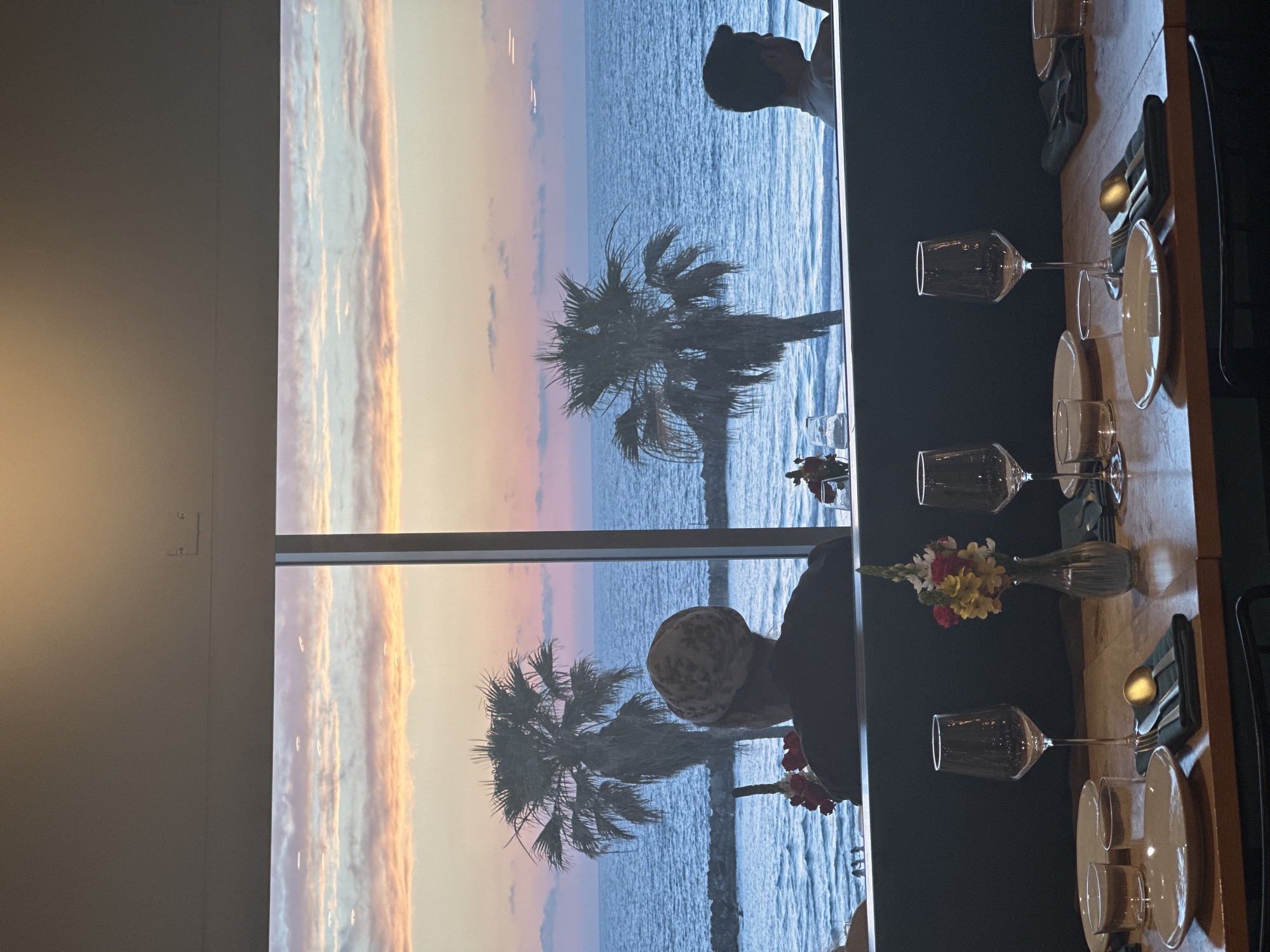 The image shows a window with a view of a Tel-Aviv beach at sunset. The sky is a blend of orange, pink, and blue hues. A palm tree is in the center of the view, silhouetted against the sky and ocean. Reflected in the window is a table setting inside what appears to be a restaurant. The table is set with plates, glasses, silverware, and napkins. A small flower arrangement sits on the table. Two people can be seen, one at the top and one at the bottom of the window.