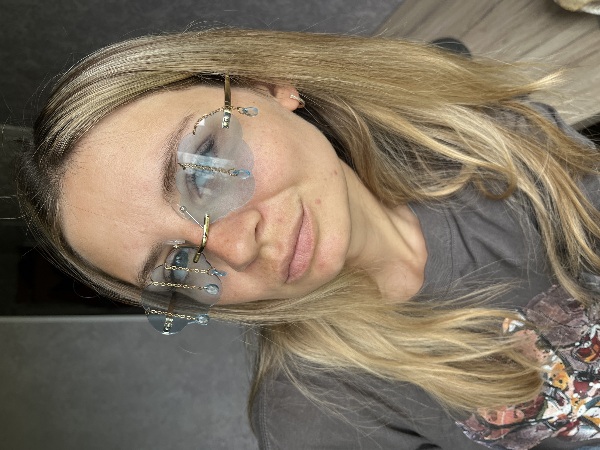 A medium shot, taken from a high angle, shows a young woman with long, blonde hair. She is lying down and her head is tilted to her left. She is wearing a grey t-shirt with a floral design on the right side of the chest. Her eyes are closed, and she is wearing unique glasses with transparent blue lenses. The lenses are shaped like clouds and are connected to a gold chain. The glasses are adorned with small, blue, teardrop-shaped gems. A gold hoop earring with small diamonds is visible in her left ear. The background appears to be an indoor setting, with a wooden surface visible in the upper right corner. The lighting suggests it could be daytime, but the overall atmosphere is casual and relaxed. No text is visible in the image.