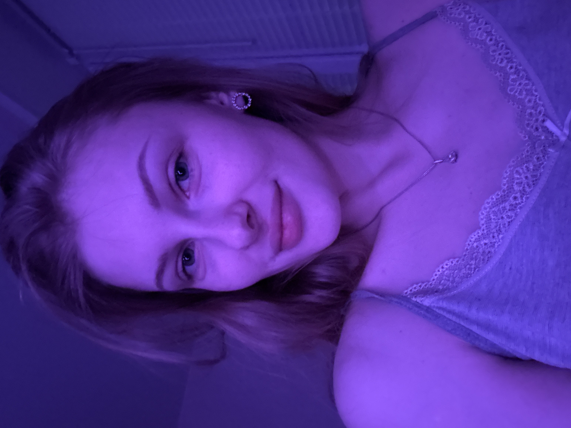 The image depicts a close-up, head-and-shoulders portrait of a young adult female, illuminated by pervasive purple lighting. She possesses fair skin, light-colored irises, and light-brown hair, styled with visible waves. Her expression is characterized by a slight, closed-mouth smile and direct eye contact with the viewer. Freckles are visible across her nose and upper cheeks. She is attired in a grey, lace-trimmed tank top. Accessories include a circular stud earring, encrusted with small reflective elements, on her proper left ear, and a slender, silver-toned necklace with a single teardrop-shaped pendant centered on her sternum. The background, largely out of focus, consists of an undifferentiated purple surface on the left and vertical slat patterns, consistent with window blinds, on the right. The precise location within Perm, Russia, cannot be determined from the visual evidence presented.