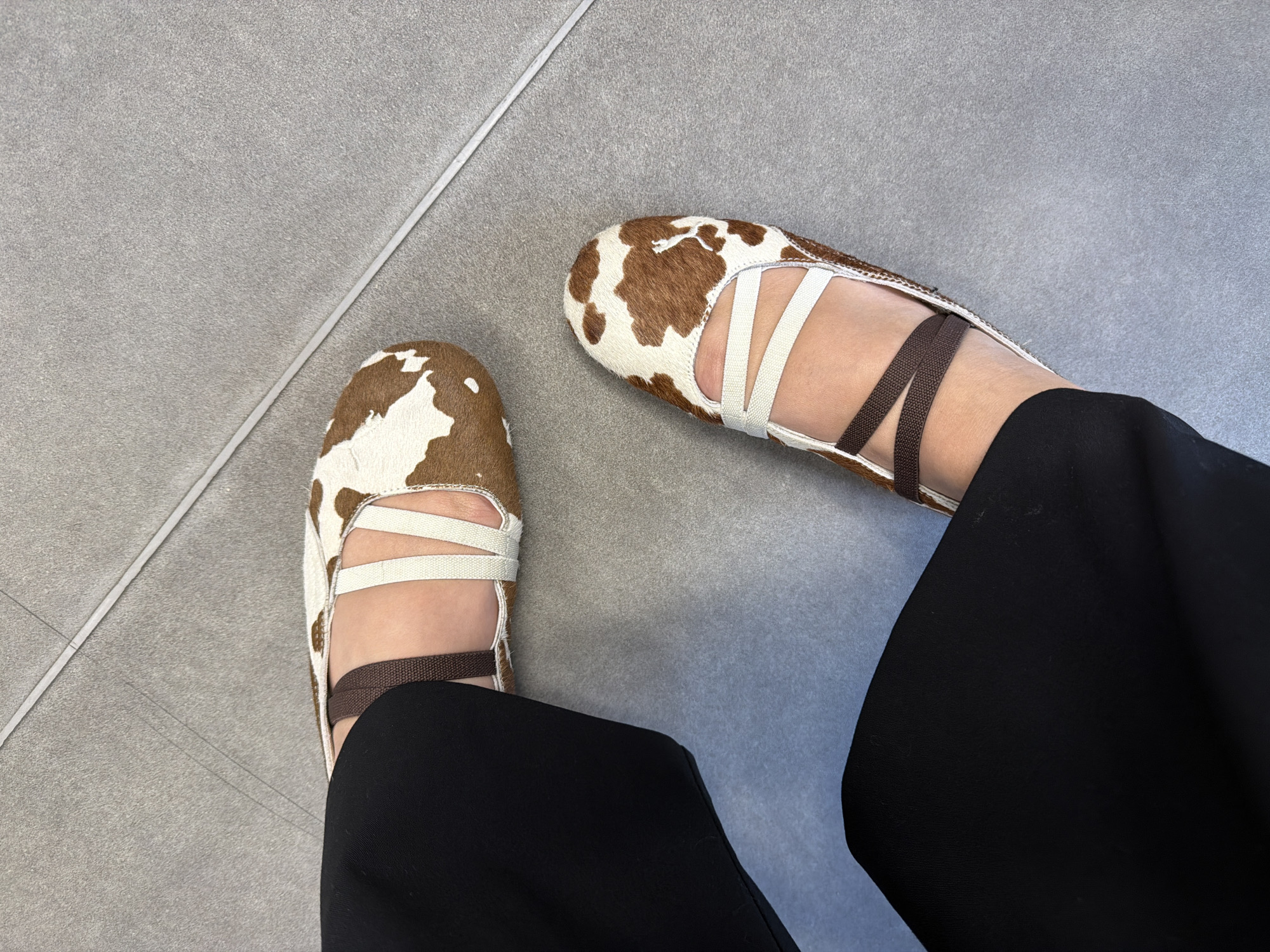 The image presents a top-down view of a person's feet clad in distinctive cow-print shoes with white and brown elastic straps. The shoes are positioned on a light gray tiled surface, and the person is wearing black pants. The background is uniform and provides no specific clues about the location. Therefore, it is not possible to determine that the location is Yerevan, Armenia from the provided image.