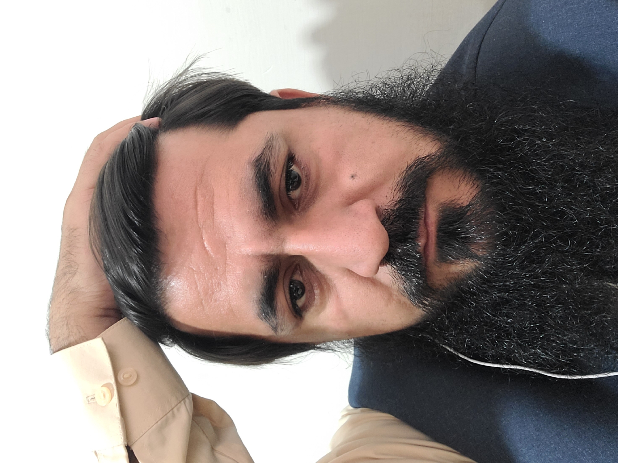 A headshot of a man with dark hair and a full black beard is shown. His right hand is resting on his head near the temple. He is wearing a light beige shirt with a button visible on the cuff, and a dark navy blue vest. The background is a plain white wall. The image does not provide any information about Quetta City Tehsil, Pakistan.