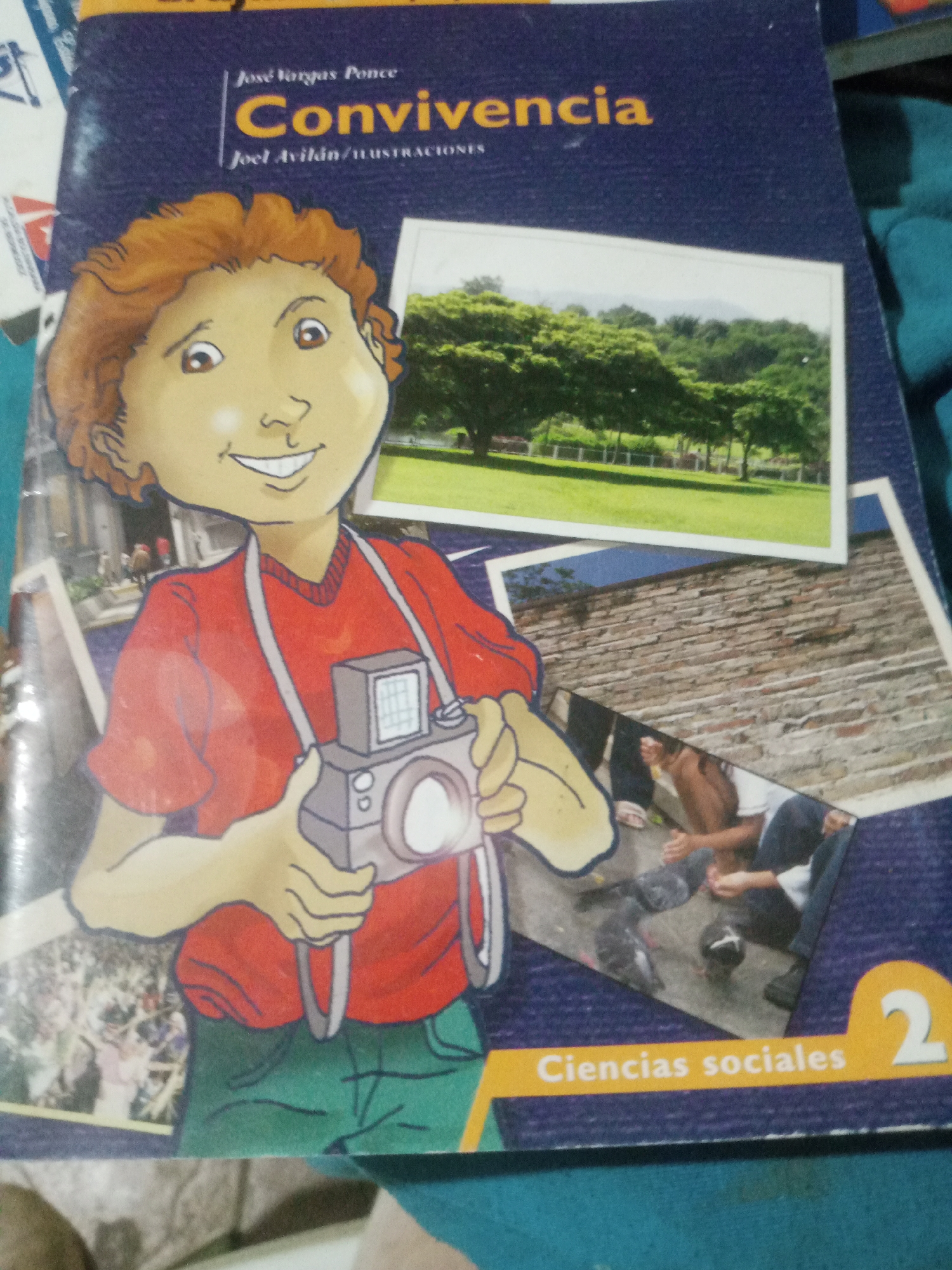 The image shows the cover of a book titled "Convivencia" by José Vargas Ponce, with illustrations by Joel Avilán. The cover features a cartoon drawing of a boy with orange hair wearing a red shirt and green pants, holding a camera. Behind the boy, there are several photographic inserts: one of a park with trees and a distant landscape, another of a brick wall, and a third, smaller one depicting people in an outdoor setting with pigeons. The bottom of the cover has a yellow banner with the text "Ciencias sociales 2" in white letters.
There is no information within the image that suggests a specific location in the city of Santa Ana, Venezuela. The content appears to be generic educational material.
