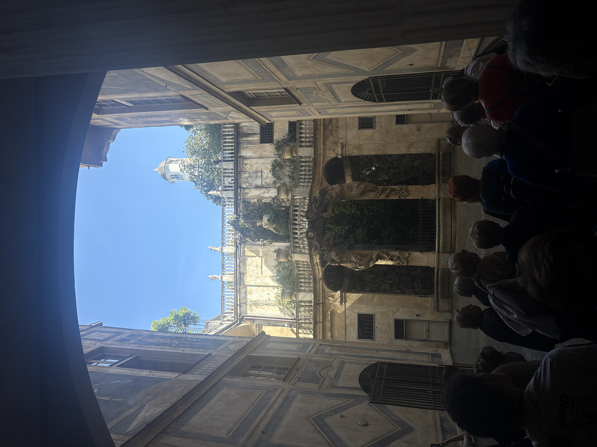 Captured from a low angle and oriented vertically, the image depicts a courtyard scene in Genoa, Italy. The architectural style is baroque. The buildings are adorned with pale walls, embellished with grey accents. There are rows of windows. There are statues and greenery. A blue sky is visible above the buildings. In the foreground, several people stand in the shadows, presumably tourists.
