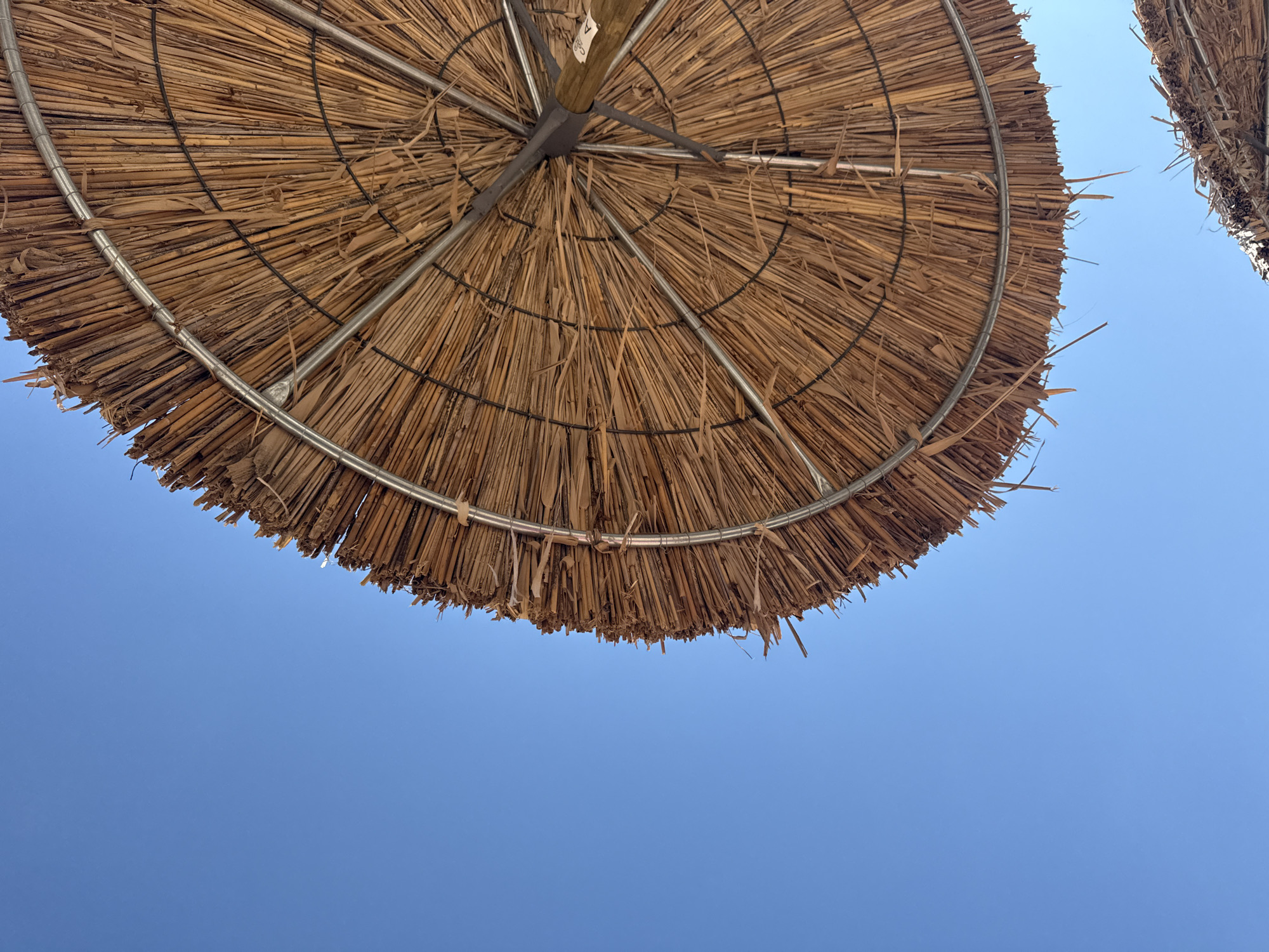 The image shows a low-angle view of a thatched umbrella against a clear blue sky. The umbrella is made of dried reeds and has a metal frame. Some loose reeds hang from the edge of the umbrella. The umbrella is located on Ofrynio Beach, Greece. The bottom right corner of the image shows a portion of another thatched umbrella.