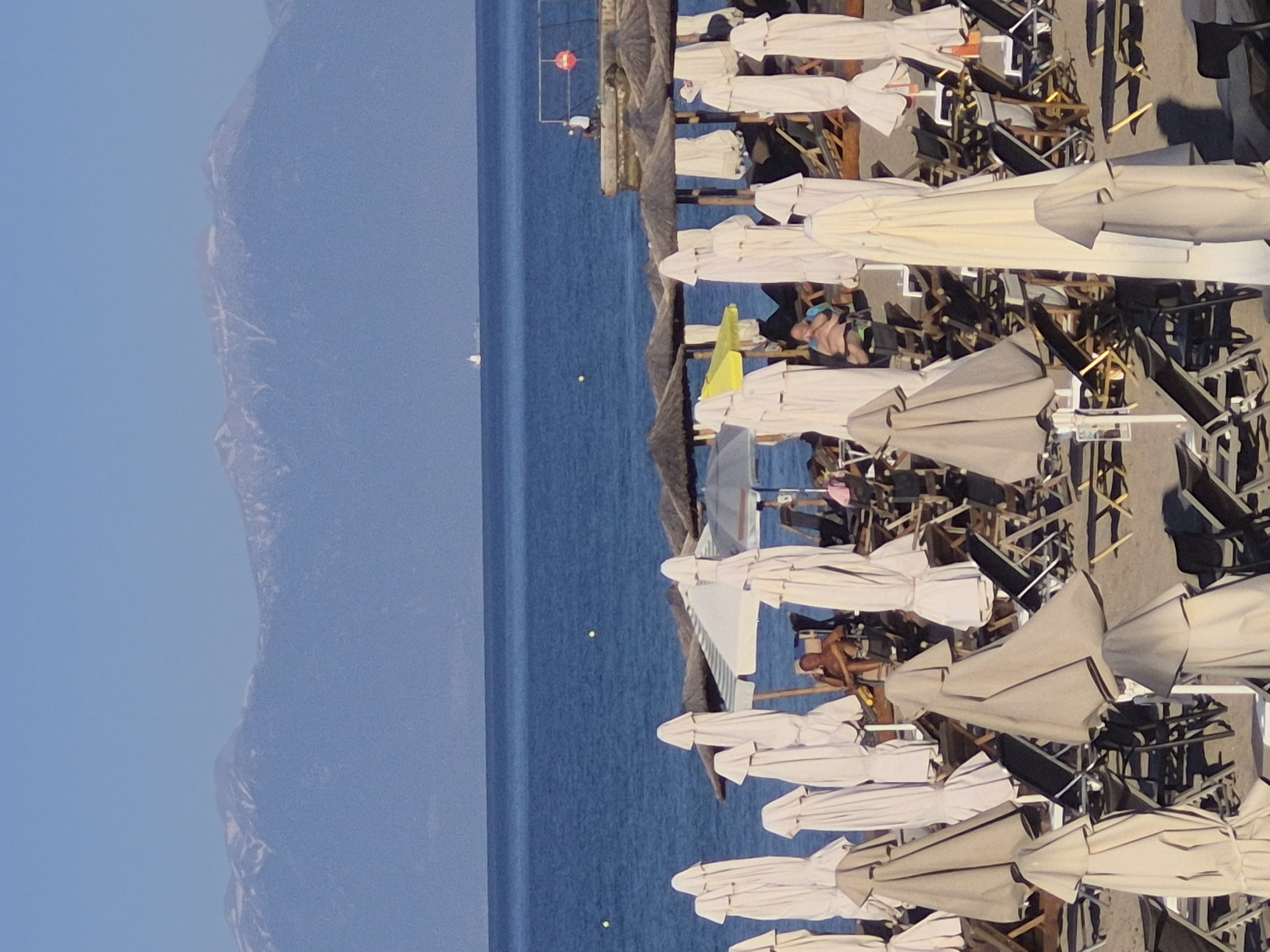 The image shows a beach scene in Νέα Καλλικράτεια, Greece. In the foreground, numerous closed beach umbrellas and lounge chairs are arranged on the sand. Some people are visible relaxing on lounge chairs. In the middle ground, the blue sea stretches out to the horizon. In the distance, mountains with snow-capped peaks are visible under a clear blue sky. A ship can be seen on the water.