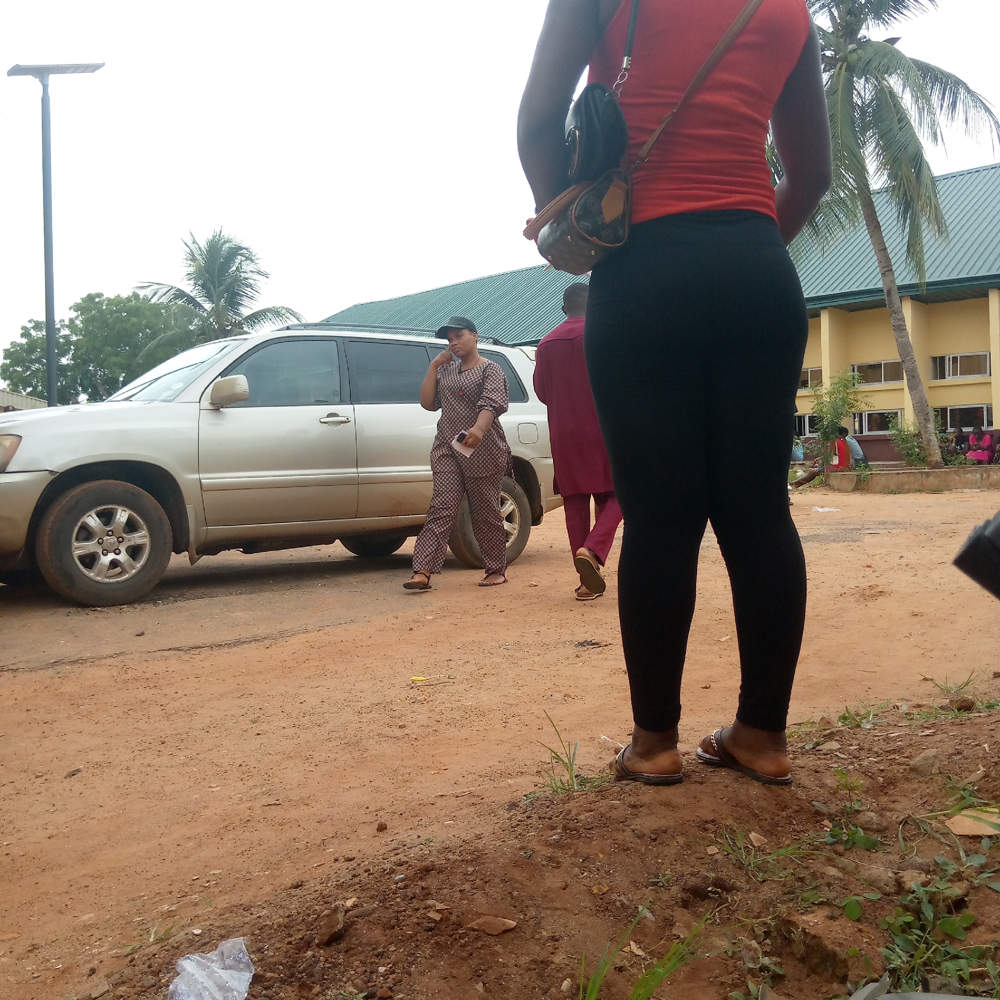 A dusty, unpaved area in Ukwu-Abwa, Nigeria shows a beige Toyota Highlander parked on the left. A woman in a patterned top and pants, with a dark cap, is walking near the car, holding a phone to her ear. Behind her, a man in a maroon outfit walks in the same direction. In the foreground, the back of a woman wearing a red top and black leggings is visible, with a purse on her shoulder. Behind the woman in red, a yellow building with a green roof is visible, and there are people sitting on a bench in front of it. A few palm trees are scattered throughout the scene. The ground is dirt and dust, with some sparse vegetation near the foreground. A light pole is visible in the upper left corner.