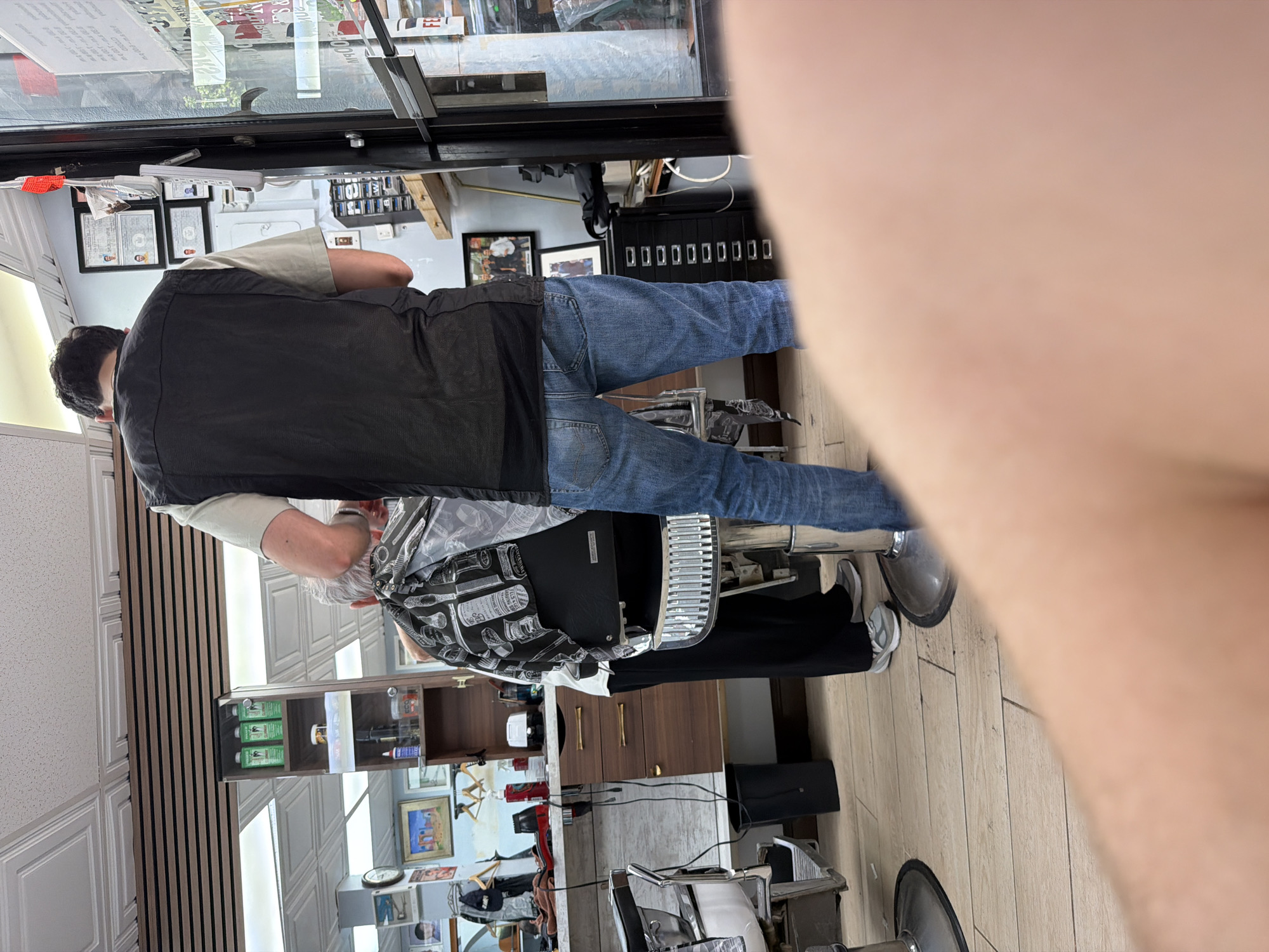 The image captures the interior of a barbershop. A person wearing a dark vest and jeans stands near a barber chair. The shop features typical barbering equipment, and shelves stocked with grooming products. Pictures and certificates are on the walls. The presence of these items, along with the distinct style of the shop, suggests this is likely in New York City, United States of America.