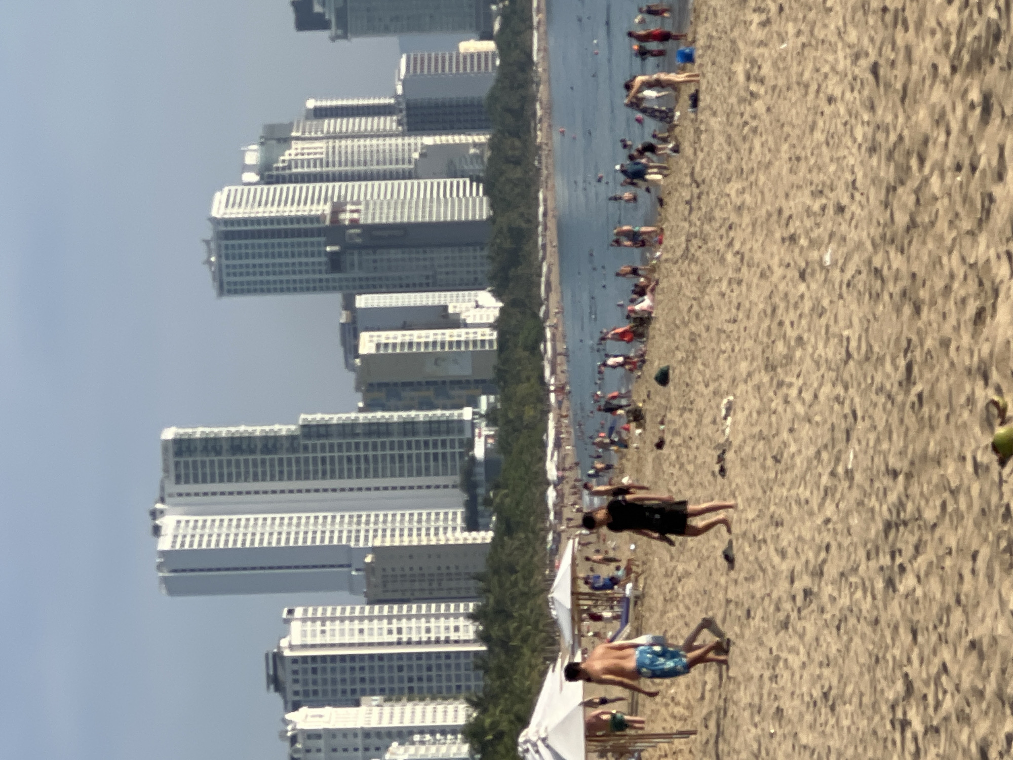 The image depicts a crowded beach in Nha Trang, Vietnam, with numerous people enjoying the water and sand. In the foreground, two men are walking on the beach, one wearing blue patterned swim trunks and the other in dark shorts. Numerous other beachgoers are visible in the middle ground and background, with many in the water and some relaxing on the sand. White umbrellas provide shade along the shoreline. Towering skyscrapers form a backdrop to the scene, indicative of a city beach environment. The water is calm, and the sky is clear with some haze. The overall scene is one of a lively summer day at a popular tourist destination.