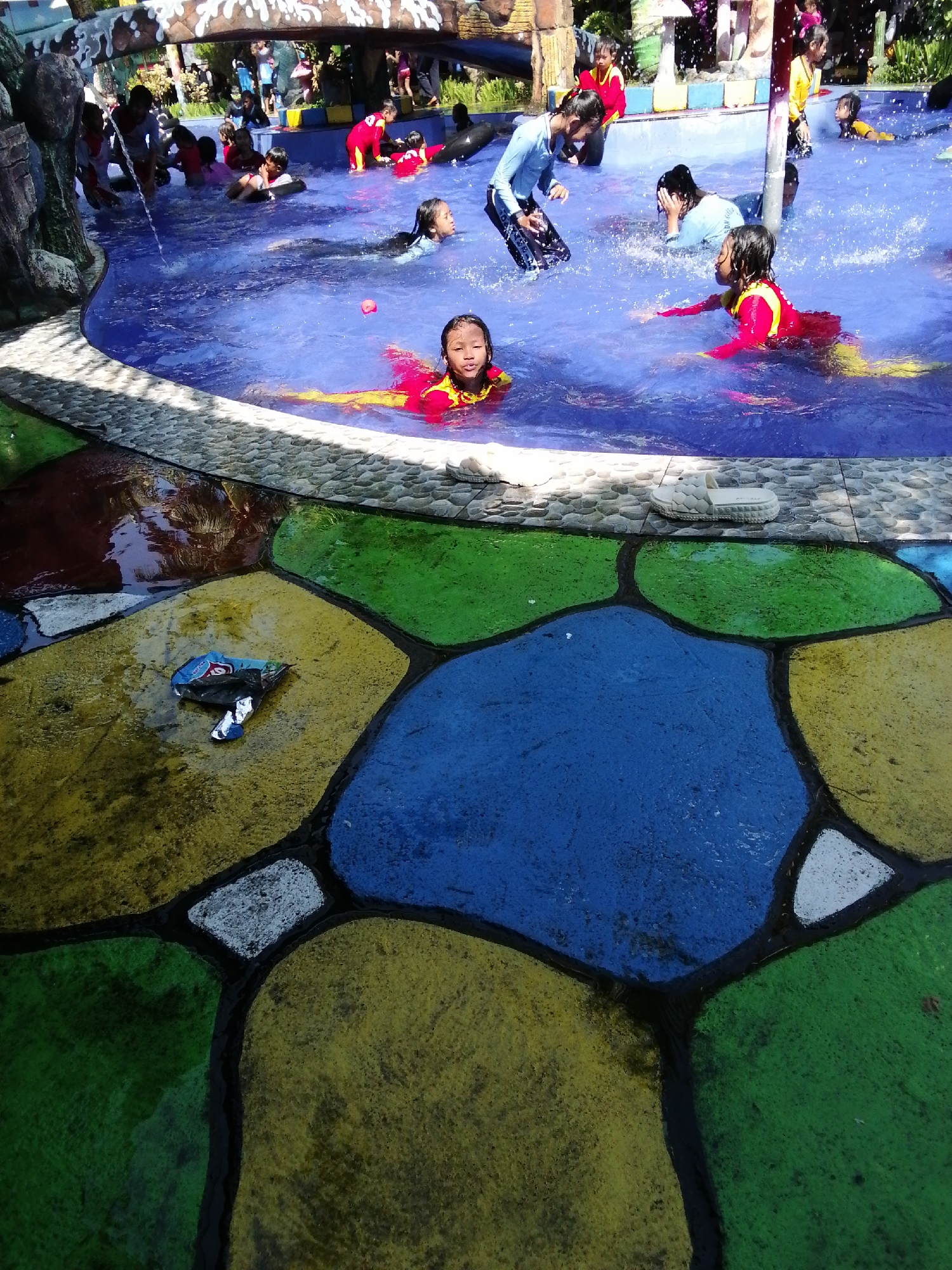 In Tegalwangi, Indonesia, at a water park, a group of children are seen playing in a pool. The pool has a blue surface with a colorful mosaic design on the surrounding pavement, featuring shapes in yellow, green, blue, and white. In the foreground, a young girl in a red and yellow swimsuit floats on her back, looking towards the camera. Behind her, another girl in a similar swimsuit is partially submerged, her head visible above the water. Further back, a group of children are engaged in various activities in the pool, some splashing water and others swimming. One child, wearing a blue long-sleeved shirt and black shorts, stands in the water with hands on their hips. The pool area features some decorative elements, including what appears to be a rock formation on the left side and a small waterfall or water feature in the background. Trash is visible on the colorful pavement near the pool's edge.