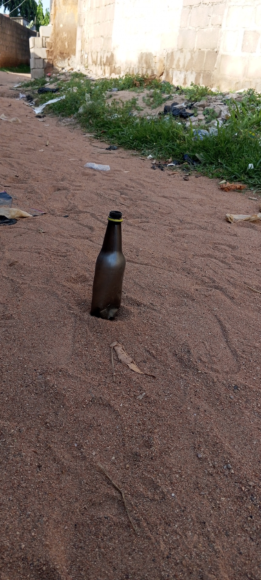 This media file depicts an outdoor scene in Jalingo, Nigeria, with a focus on a brown plastic bottle lying on reddish-brown sand. The bottle is empty and uncapped, standing upright and slightly tilted. A thin yellow band is visible around the neck of the bottle.
The setting appears to be a dusty area, possibly a roadside or an undeveloped plot of land. In the background, a building constructed with light-colored concrete blocks is visible. The wall shows signs of wear and some staining. Adjacent to the building, there is a patch of green vegetation, interspersed with some debris, including black plastic bags and white plastic wrappers, suggesting an area with litter.
Further to the left of the building, another wall, constructed with grey concrete blocks, is visible. The ground is predominantly sandy, with some small stones and dried leaves scattered across it. A single dry leaf is prominently positioned near the base of the bottle. A thin, dry grass stalk lies on the sand to the right of the bottle.
The lighting suggests it is daytime, possibly late afternoon, as the sunlight casts some subtle shadows. There are no people visible in the frame, and the overall impression is one of a somewhat neglected or undeveloped urban or semi-urban environment. No text is visible in the image.
