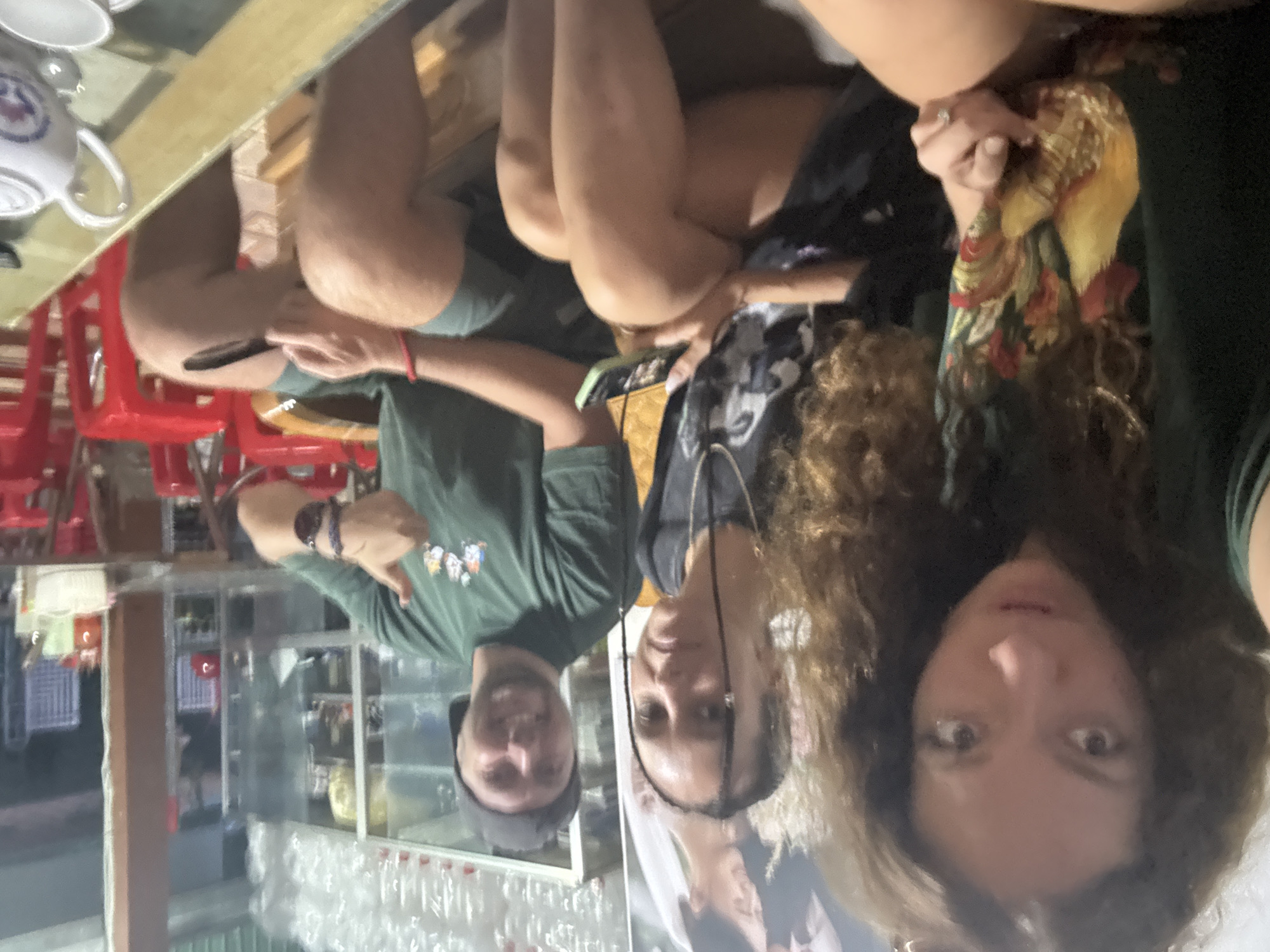 Three individuals, two women and one man, are captured in a group photo or selfie, apparently taken indoors in Mù Cang Chải, Vietnam. The image is presented rotated 90 degrees clockwise from its likely original orientation, so subjects appear to be on their sides.
The man is positioned on the left side of the frame (when corrected), wearing a dark green t-shirt and a dark baseball cap. He has a beard and is smiling directly at the camera, gesturing with his left thumb. His left arm also shows a red bracelet.
Next to him, in the center, is a woman with dark, braided hair, looking towards the camera with a neutral expression. She is wearing a dark top.
On the right side of the frame is another woman with voluminous curly brown hair, looking at the camera with wide eyes. She is wearing a dark top and holds a patterned scarf or piece of fabric in shades of yellow, red, and green.
In the foreground, a light-colored wooden table is visible, on which a white ceramic teapot with a blue design and a matching cup are placed. Red plastic chairs are partially visible beneath the table. The background features the interior of what appears to be a shop or cafe, with shelves displaying various items, possibly bottles or other goods, behind glass panels that reflect parts of the room. The scene is illuminated by indoor lighting, suggesting it might be daytime. No discernible text is visible. The atmosphere appears casual and interactive as the subjects pose for the photo.