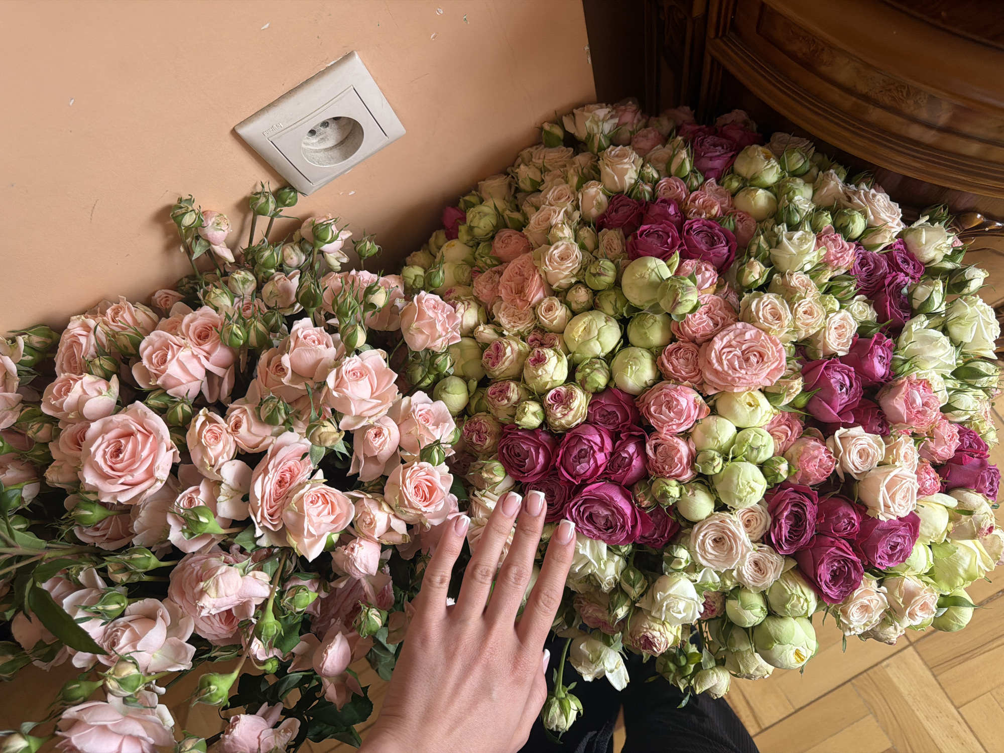 The image shows a floral arrangement with a hand placed over it, set indoors. A large bouquet of mixed roses in shades of pink, white, and deep magenta fills the right half of the frame, extending towards the top right corner where a wooden cabinet is partially visible. The left half features a separate arrangement of pale pink roses. The hand, with manicured nails, rests on these pink roses. In the background on the left is a peach-colored wall with a white electrical outlet. The location is likely an interior space, but without additional context, the city or the country of Yerevan, Armenia, cannot be confirmed.