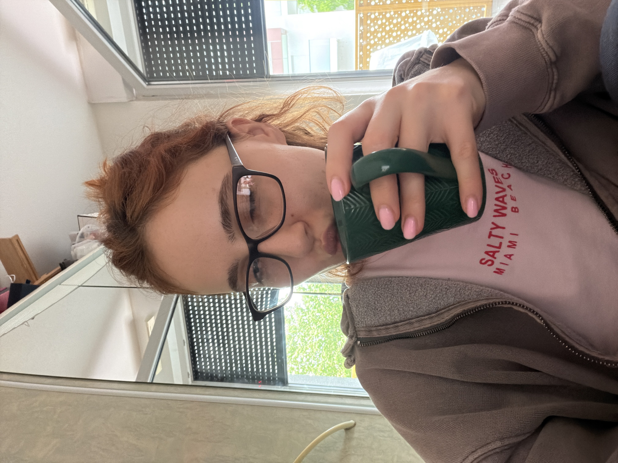 A young woman with auburn hair pulled up, wearing black-rimmed glasses and a light pink t-shirt with red lettering that reads "SALTY WAVES MIAMI BEACH", is holding a dark green mug with both hands and sipping from it. She is also wearing a light brown zip-up hoodie. The background shows a window with a roller blind, revealing lush green foliage outside, suggesting a daytime setting. The woman's fingernails are painted light pink. The environment appears to be indoors, possibly a home or cafe, with a mirror reflecting part of the room. The image is taken from a close-up perspective, focusing on the woman's face and upper body. There is no specific information in the image that indicates the location being Strasbourg, France.