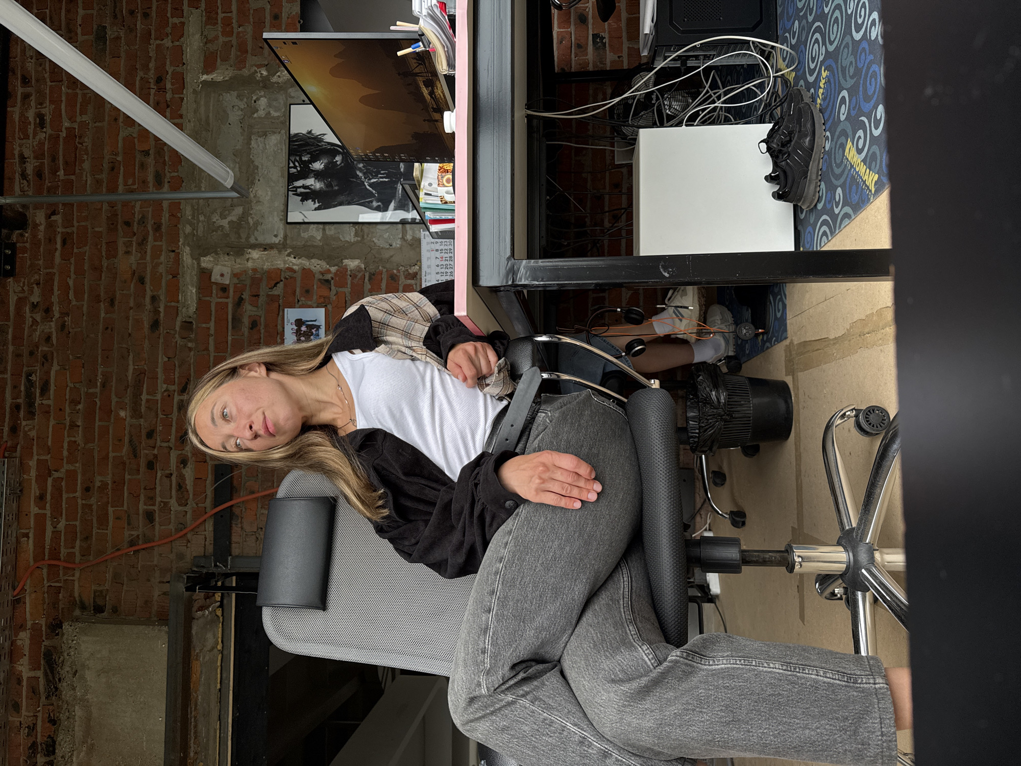 A young woman with blonde hair is sitting in an office chair with her legs crossed. She is wearing a white tank top, a black jacket, and grey jeans. She is looking to the side and appears to be deep in thought. The office is decorated with exposed brick walls and has a desk with a computer, various office supplies, and a black trash can. A pair of black sneakers are on the floor next to the desk. The location is not specified in the image or the prompt.