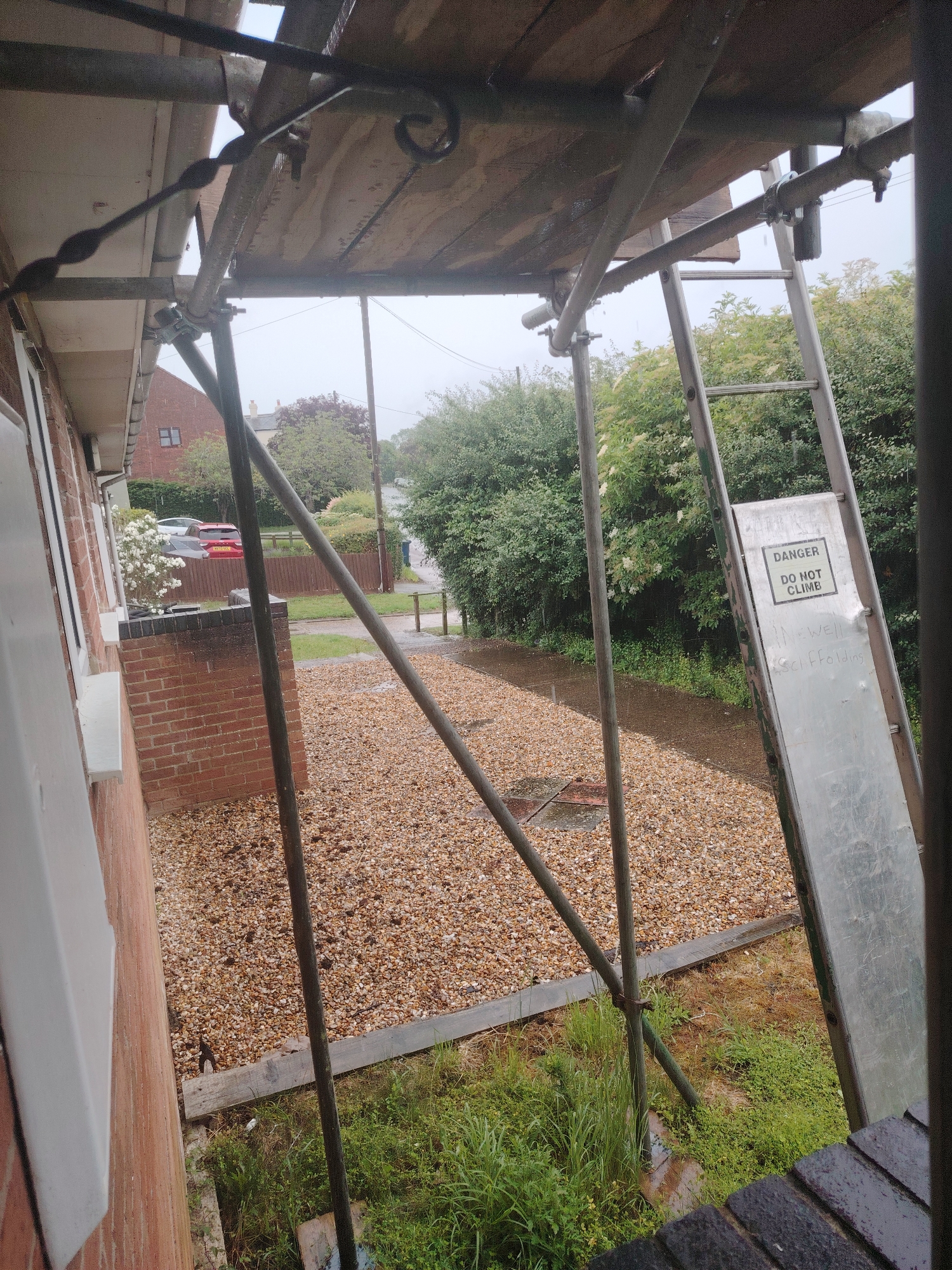 An outdoor, slightly angled shot shows scaffolding erected alongside a brick house in Huntingdonshire, United Kingdom. The scaffolding is made of metal poles and wooden planks, with a ladder attached to one side. A "DANGER DO NOT CLIMB" sign is affixed to the ladder. The ground is covered with gravel and some patches of grass and weeds. In the background, trees and a street can be seen, along with two parked cars. The weather appears overcast, possibly raining, as evidenced by the wet surfaces.