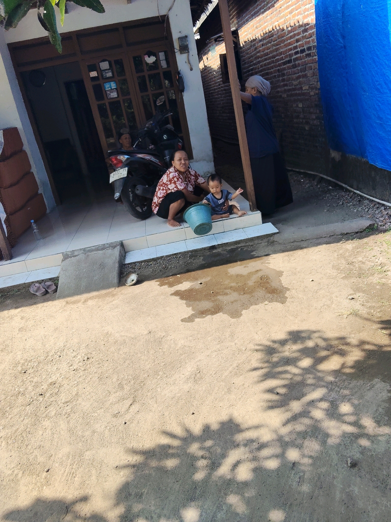 In Windurejo, Indonesia, a scene unfolds outside a home. In the foreground, a sandy, uneven surface is dappled with the shadows of overhead foliage. To the left, a concrete ramp leads to a tiled porch. A pair of brown flip-flops rests on the ground near the ramp. A motorcycle, license plate S 6122 PX, is parked on the porch.
A woman, wearing a patterned red and white blouse and black shorts, squats on the porch, holding a light blue bucket. Beside her, a young child, dressed in a blue shirt with the word "COOL" on it, sits on the tiled steps, looking towards the right. Further back, another person, partially obscured, sits on the motorcycle.
To the right of the porch, a brick wall rises, partially covered by a large blue tarp. A woman wearing a blue shirt and a light-colored headscarf stands near a wooden post, looking towards the wall. A bottle of water is visible on the tiled surface near the motorcycle. The overall environment suggests a casual, domestic setting.