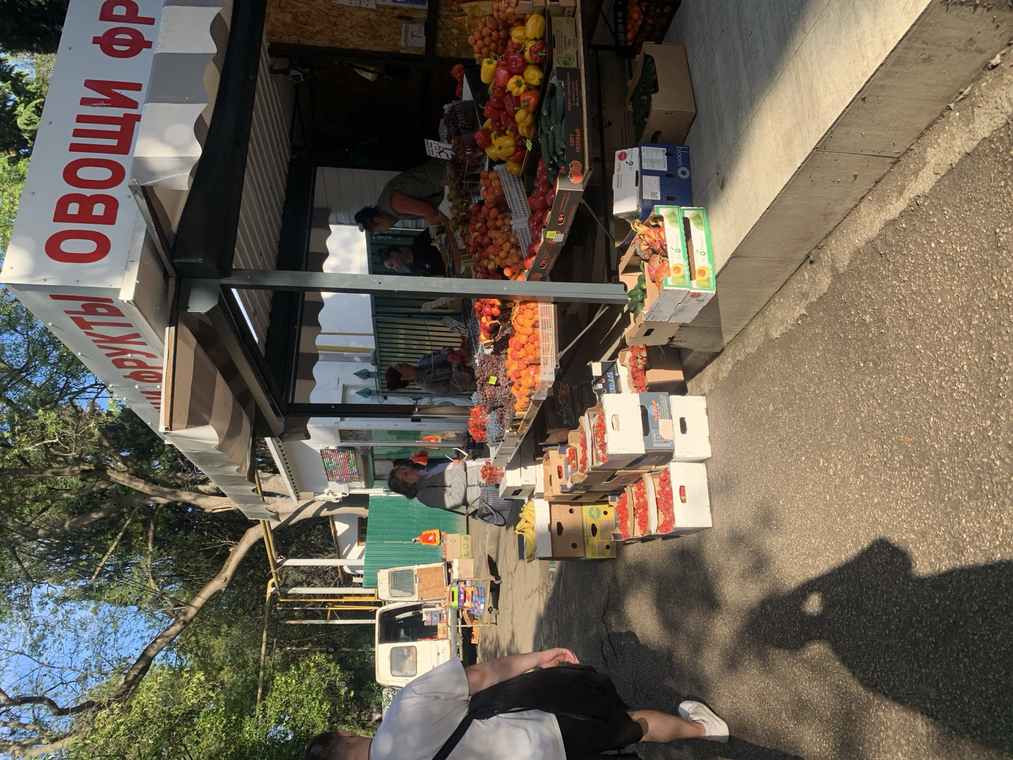 The image captures a street-level view of a fruit and vegetable stand. The stand has a sign in Cyrillic script reading "FRUITS VEGETABLES." A variety of produce is displayed, including peppers, tomatoes, cucumbers, grapes, and other fruits. People are both manning the stand and appearing to be customers. Boxes of strawberries are stacked on the ground near the stall. A parked car with a trailer is slightly in the background. The lighting indicates it is daytime. There are trees on the left. The location is in the city of Unknown, Unknown.