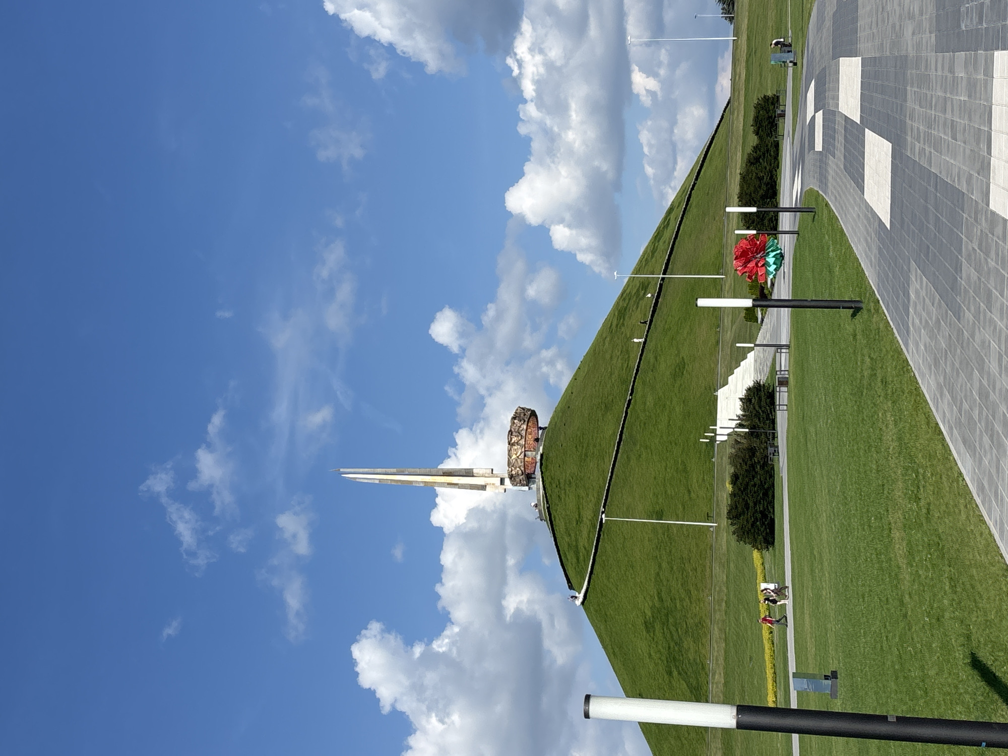 The image depicts the mound of glory in the city of Ozyaritska-Slabadski rural council, Belarus, under a bright blue sky with scattered white clouds. The mound is covered in neatly trimmed green grass, with a winding pathway leading to the summit where a tall obelisk-shaped monument stands. The monument is adorned with a circular structure at its base featuring reliefs and inscriptions. Several people are visible on the pathway and at the base of the mound. In the foreground, a paved walkway with a pattern of grey and white squares curves into the scene. Several modern lampposts are positioned along the pathway. To the left of the walkway, a vibrant red artificial flower sculpture is displayed between two lampposts. Green bushes are present in the middle ground, and the overall environment is clean and well-maintained, suggesting a public park or memorial site.