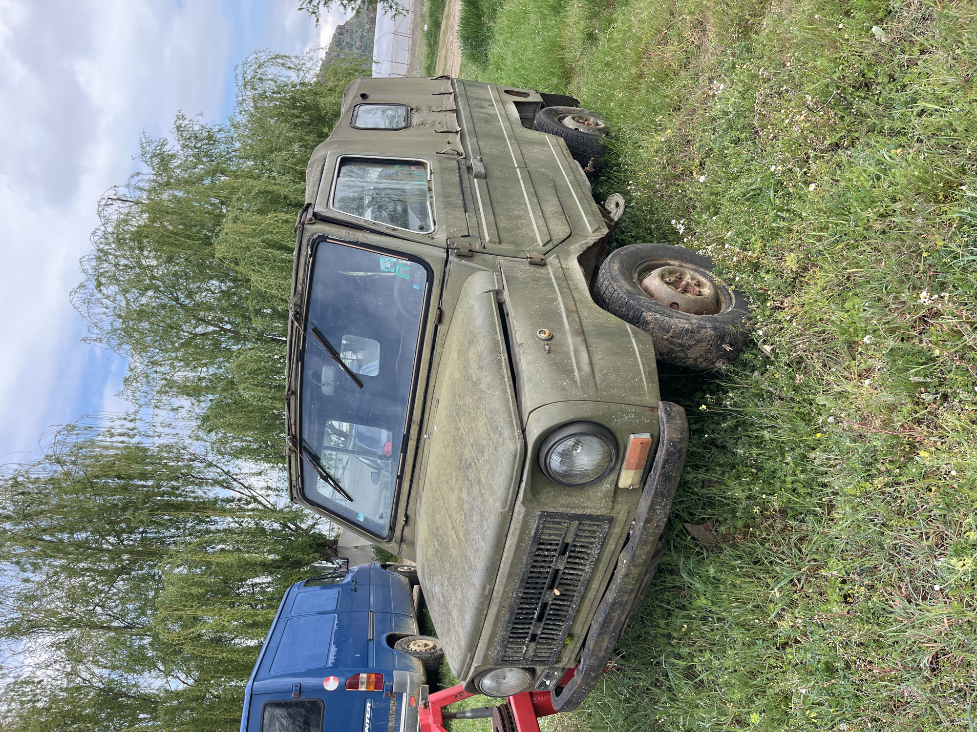 The image shows a weathered, olive-green military vehicle, possibly a LuAZ-969, parked on grassy terrain. The vehicle is partially obscured by tall grass and weeds. A blue van is visible in the background, parked nearby. The overall scene suggests a rural or semi-rural location in Lebnitsa, Bulgaria. The sky is partly cloudy. The vehicle appears neglected and possibly abandoned.