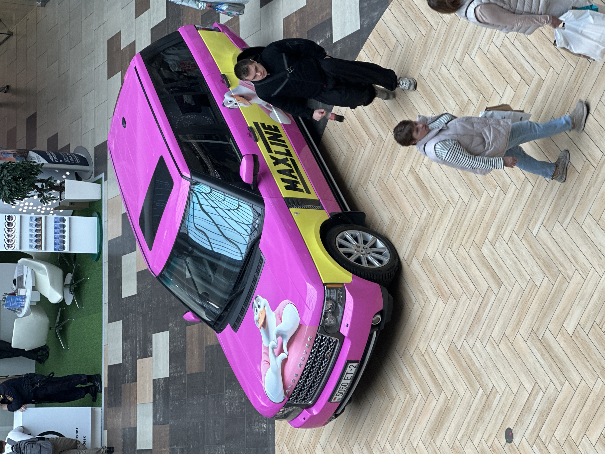 An overhead, high-angle shot captures a bright pink and yellow Range Rover parked indoors on a wood-patterned tiled floor, likely in a shopping mall. The vehicle features a cartoon duck graphic on its hood and the word "MAXLINE" emblazoned on its side. Several people are visible: a man in a black tracksuit walks beside the car, a woman in a beige vest and striped shirt walks in the foreground, and another woman in a similar vest is partially visible on the right. In the background, other individuals, seating areas, and display shelves can be observed. The car's license plate is visible and reads "BY 5050 ET-2". The overall impression is of a vibrant, commercial setting with an eye-catching vehicle as a central focus.