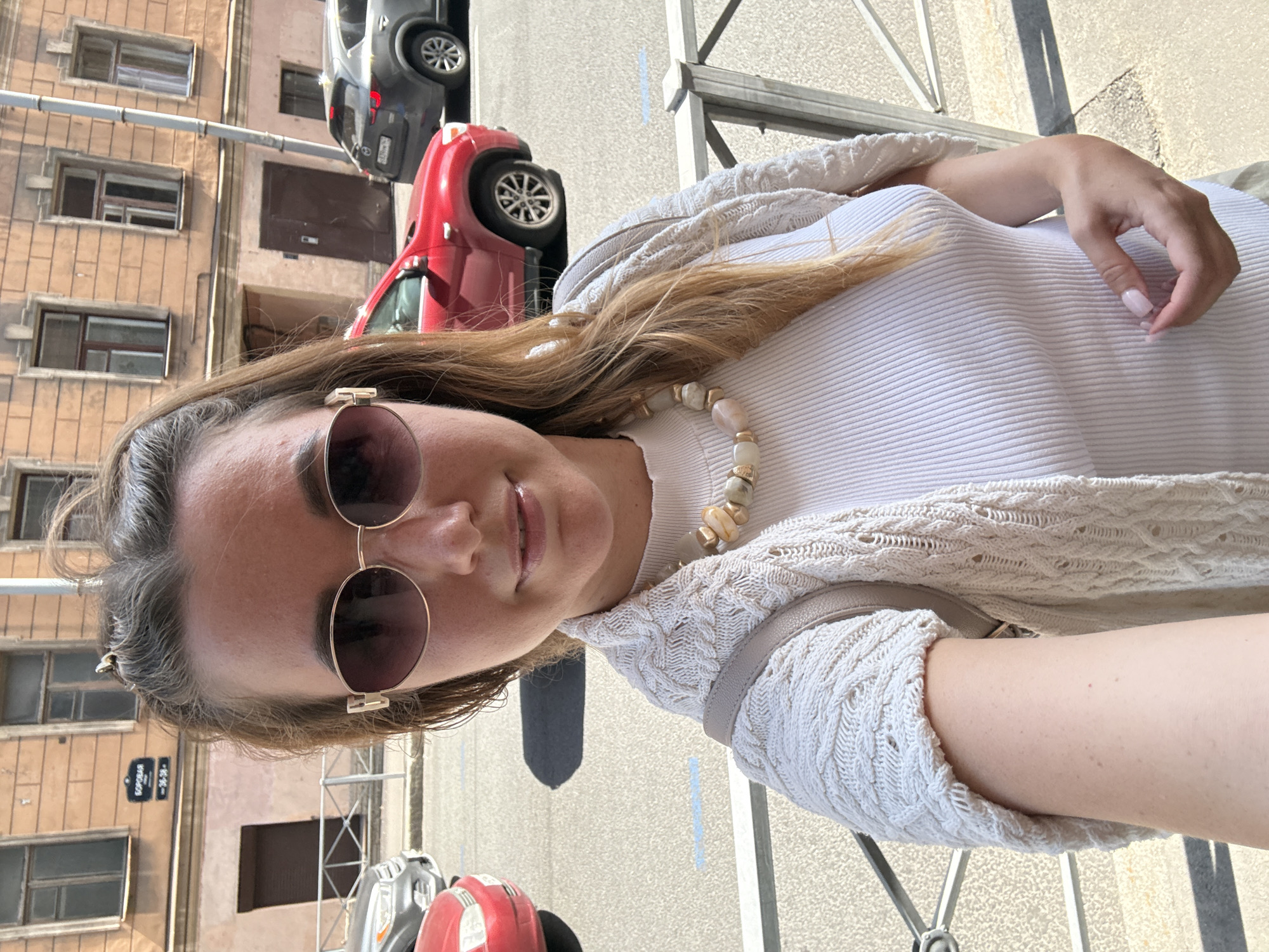 A medium-close-up selfie, rotated 90 degrees counter-clockwise, captures a woman outdoors on a sunny day. The woman, with light brown hair, is wearing aviator-style sunglasses with dark lenses. She has a slight smile on her face. She is dressed in a white, ribbed long-sleeved top and a light cream or beige knitted cardigan with intricate patterns. Around her neck, she wears a necklace made of irregular, light-colored, possibly stone or shell-like beads. Her left hand is visible, resting on her midsection, showing light pink nail polish.
The background depicts a city street scene. Behind her, there is an old, multi-story building with several windows. Parked on the street are a bright red car and a dark-colored SUV. A metal railing or fence is visible in the foreground to her right. The ground appears to be a paved sidewalk or street.
The overall impression is bright and sunny, indicating daytime. A street sign on the building behind her is visible, displaying the text "БОРОВАЯ" (Borovaya) and numbers "30-38".