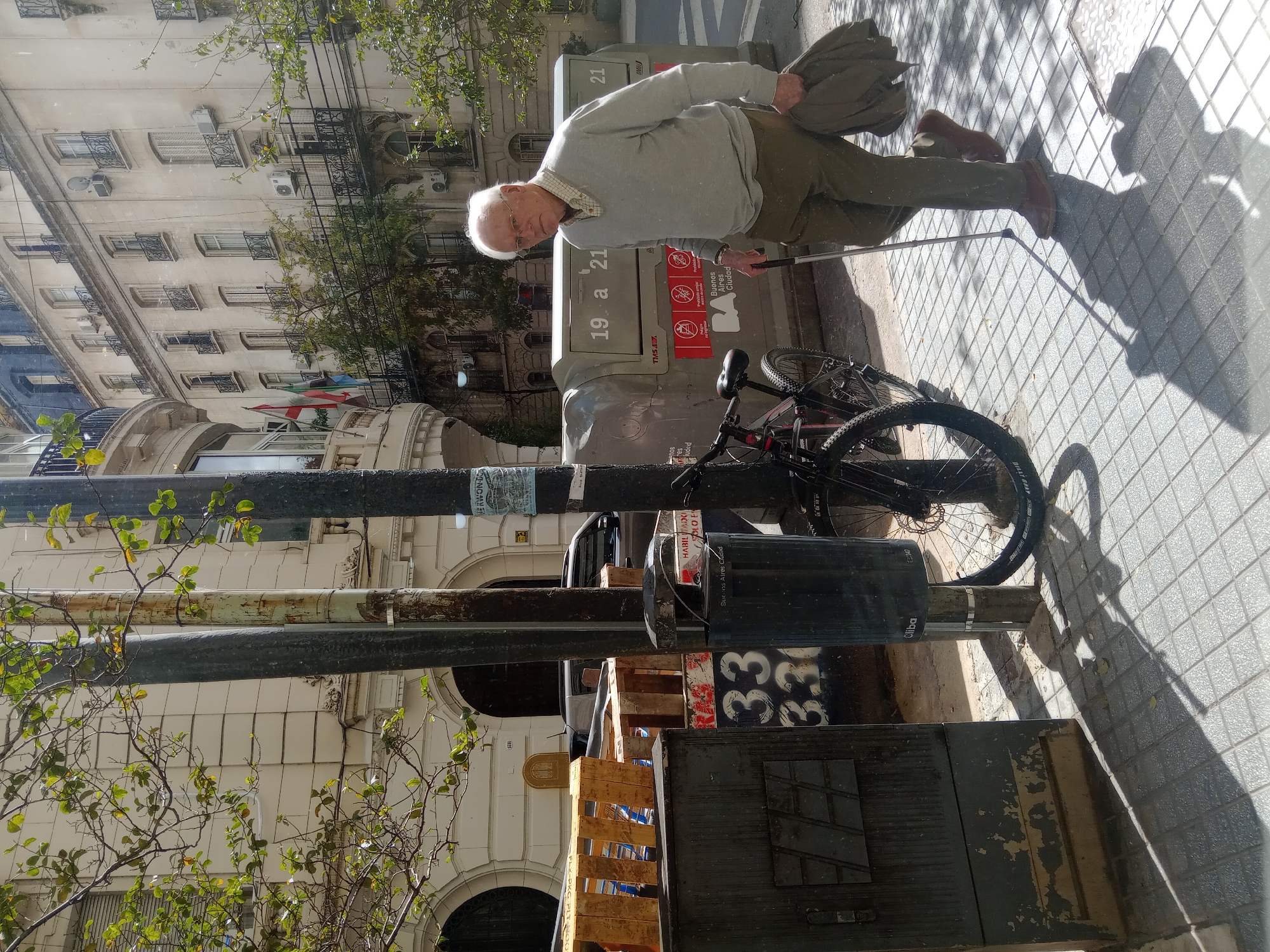 The image shows an older man standing on a tiled sidewalk in Buenos Aires, Argentina, holding a cane in his left hand and a jacket or coat in his right. He is wearing a light grey sweater and brown pants. Behind him is a modern-style bicycle parked next to a grey public recycling bin. The bin has the text "19 a 21" and "TMSAZ" printed on it, along with instructions. A dark, rectangular object with graffiti, including the number "33", is located in front of the bicycle.
The background features a building with multiple windows and architectural details. There are trees along the street, and light poles are visible. The sidewalk is shaded, indicating a time of day with strong sunlight. The sticker says "HABILITAD". The recycling bin also has a warning about improper waste disposal.