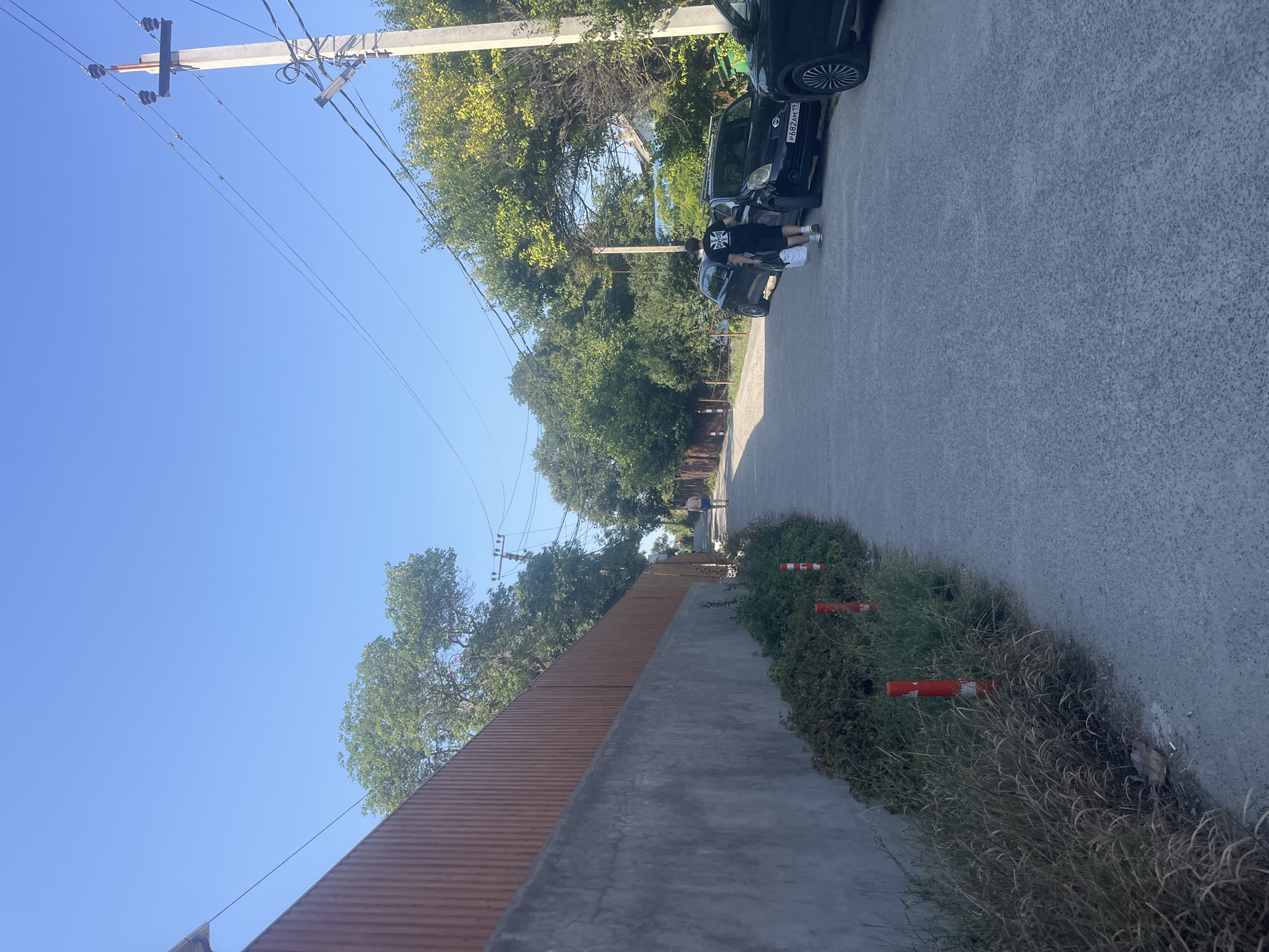 In an outdoor scene with a clear blue sky, a paved road winds uphill. On the left, a tall, ribbed brown fence with a grey concrete base runs along the side of the road, bordered by overgrown dry grass and a few red and white striped bollards. On the right, parked cars line the street. A young person wearing a black t-shirt with a graphic print and black shorts walks away from the camera, carrying two white bags. Further up the road, another person, wearing blue shorts, walks in the distance. Overhead, power lines are strung between concrete utility poles. Lush green trees and foliage are visible on both sides of the road, casting shadows.