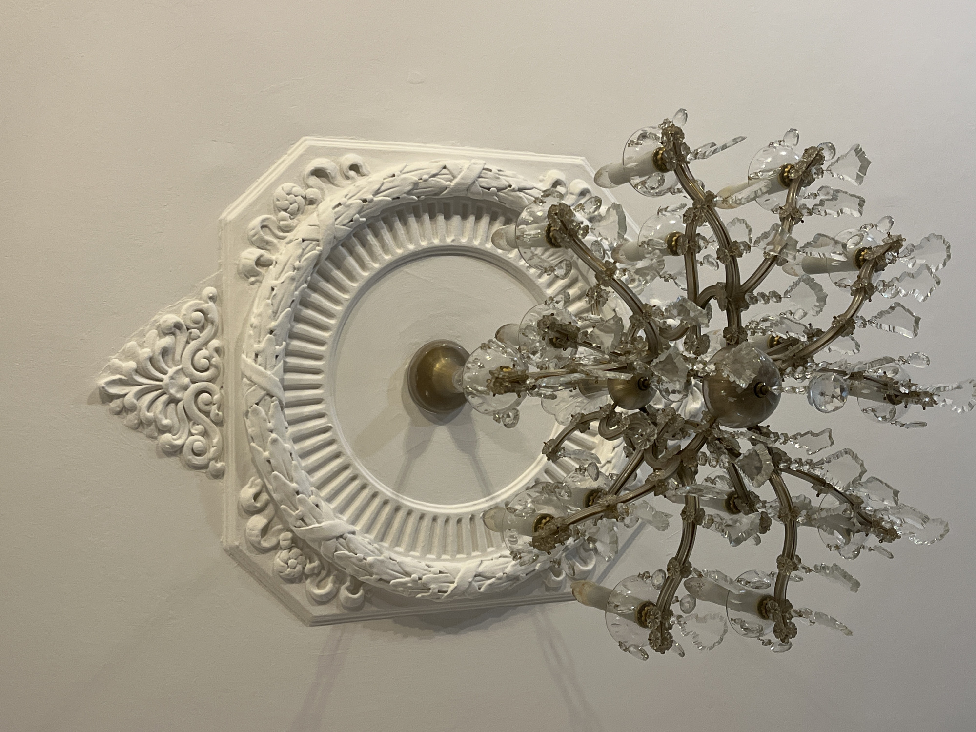 The image shows a chandelier hanging from a decorative ceiling medallion in an interior space. The ceiling is painted white. The medallion features a circular central element with concentric rings and detailed floral or leaf ornamentation on its outer edges and corners. The chandelier is ornate, with multiple arms supporting light fixtures and adorned with numerous clear crystal pendants, reflecting light. The chandelier's metal frame is likely brass or a similar golden-toned metal. The image provides no information indicating it was taken in Belgrade, Serbia.