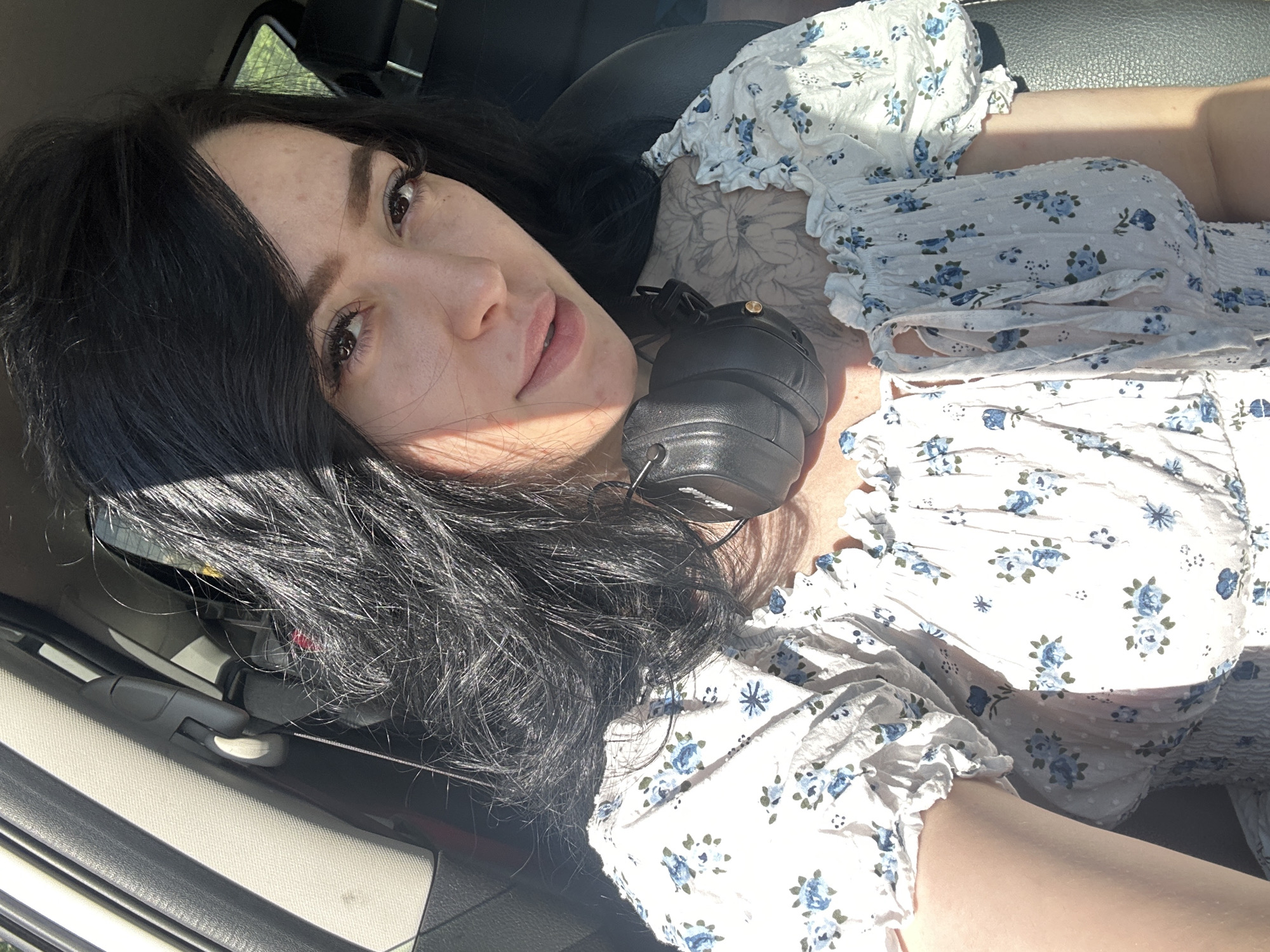 A woman with dark hair is reclining, wearing a white floral-patterned blouse, with headphones around her neck. She appears to be inside a vehicle. There is no evidence of the location of Izhevsk, Russia.