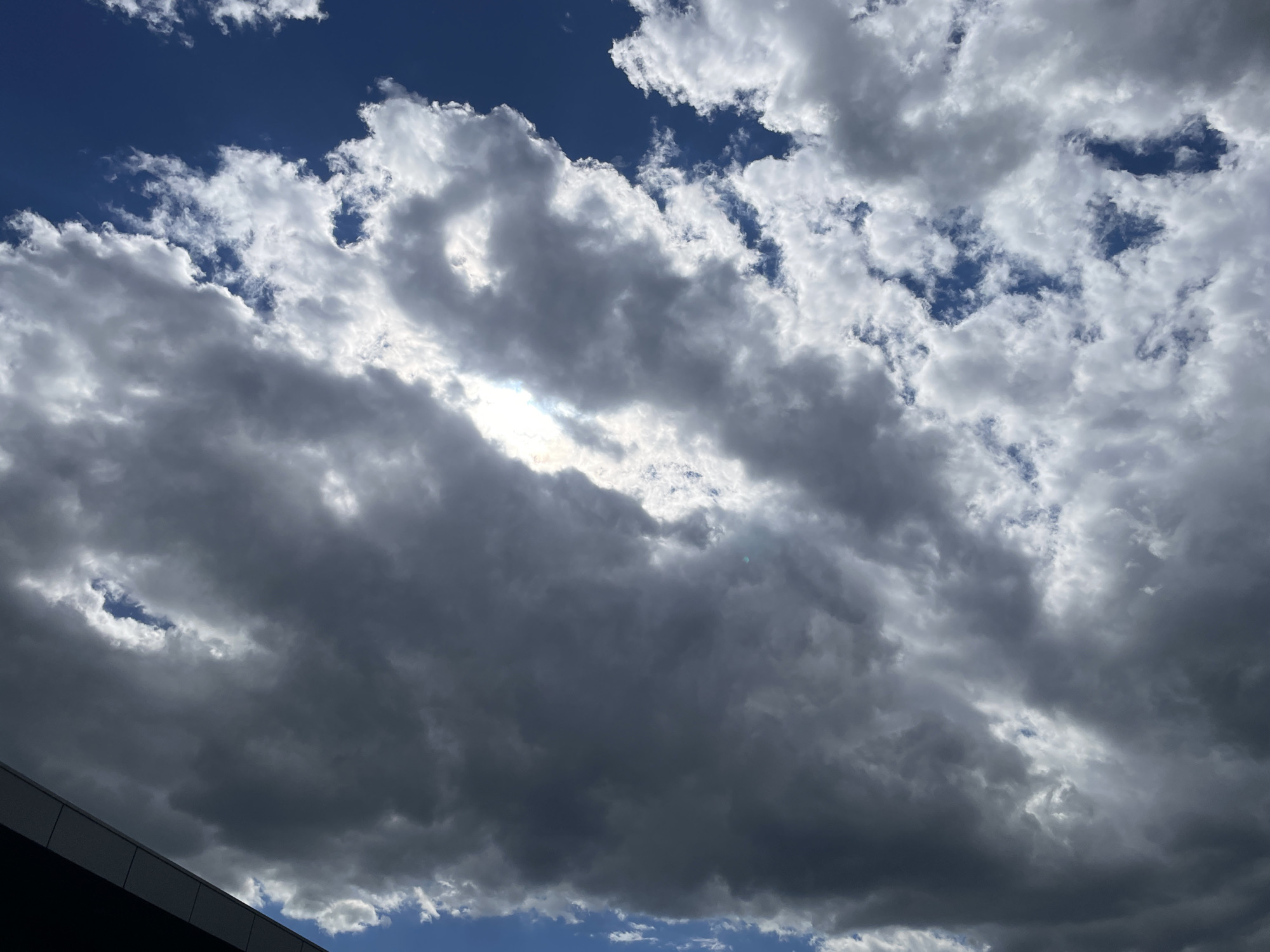 The image shows a sky filled with cumulus clouds, varying in shades of gray and white, against a backdrop of blue sky. The clouds are dense and layered, with sunlight peeking through a gap in the center. A portion of a dark-colored building is visible in the lower-left corner of the frame. The location in the city of Unknown, Unknown, is not discernible from the image.