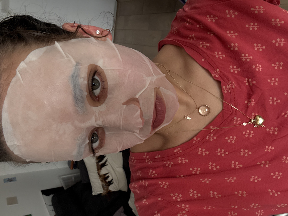 A female individual is depicted in a close-up, chest-up view, appearing to be in an indoor setting in Milan, Italy. Her face is covered by a translucent white sheet mask that adheres to her skin, leaving her dark green/hazel eyes, prominent eyelashes, eyebrows, and lips exposed. Her lips exhibit a reddish-pink tint. Dark hair is visible at the temples and behind her ears. She wears multiple piercings: a small clear stud in each earlobe and a silver-toned hoop in the helix of both ears. She is clad in a reddish-pink V-neck t-shirt patterned with small, light-colored, stylized floral or plant motifs. Around her neck, she wears two gold-toned necklaces: a shorter one with a circular, possibly iridescent, shell-like pendant and a small rectangular tag, and a longer one featuring a prominent golden heart-shaped pendant with a small white bead. The background to her left is out of focus, revealing dark furniture, a white pillow with a dark pattern, and a light-colored container. The background to her right shows a plain light-colored wall and a darker floor. The subject is directly facing the camera.