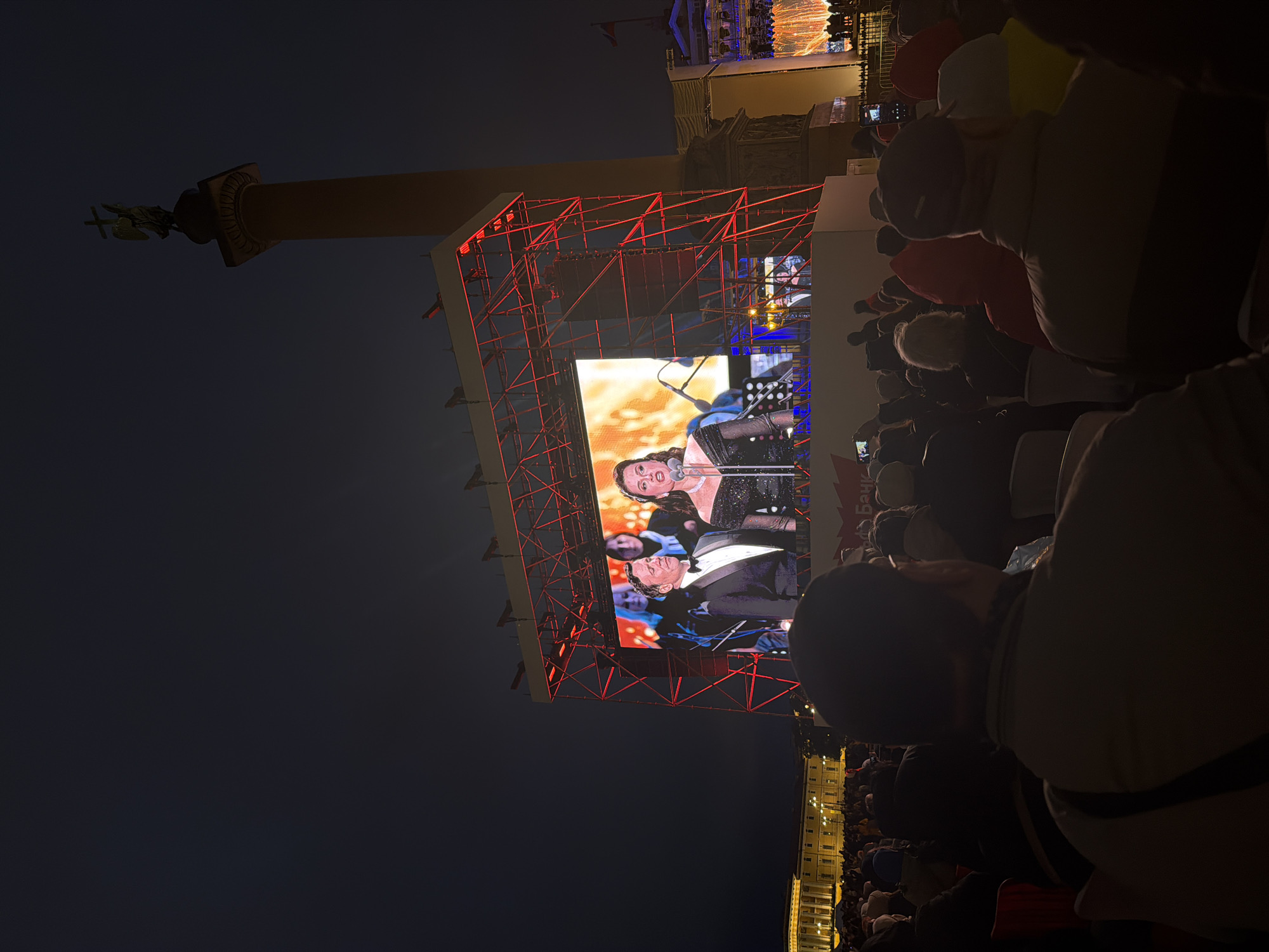 A nighttime crowd gathers in Palace Square, Saint Petersburg, Russia, to watch a large screen projecting the image of a man and woman in formal attire. The Alexander Column is visible on the left of the screen. The Winter Palace is visible in the background.
