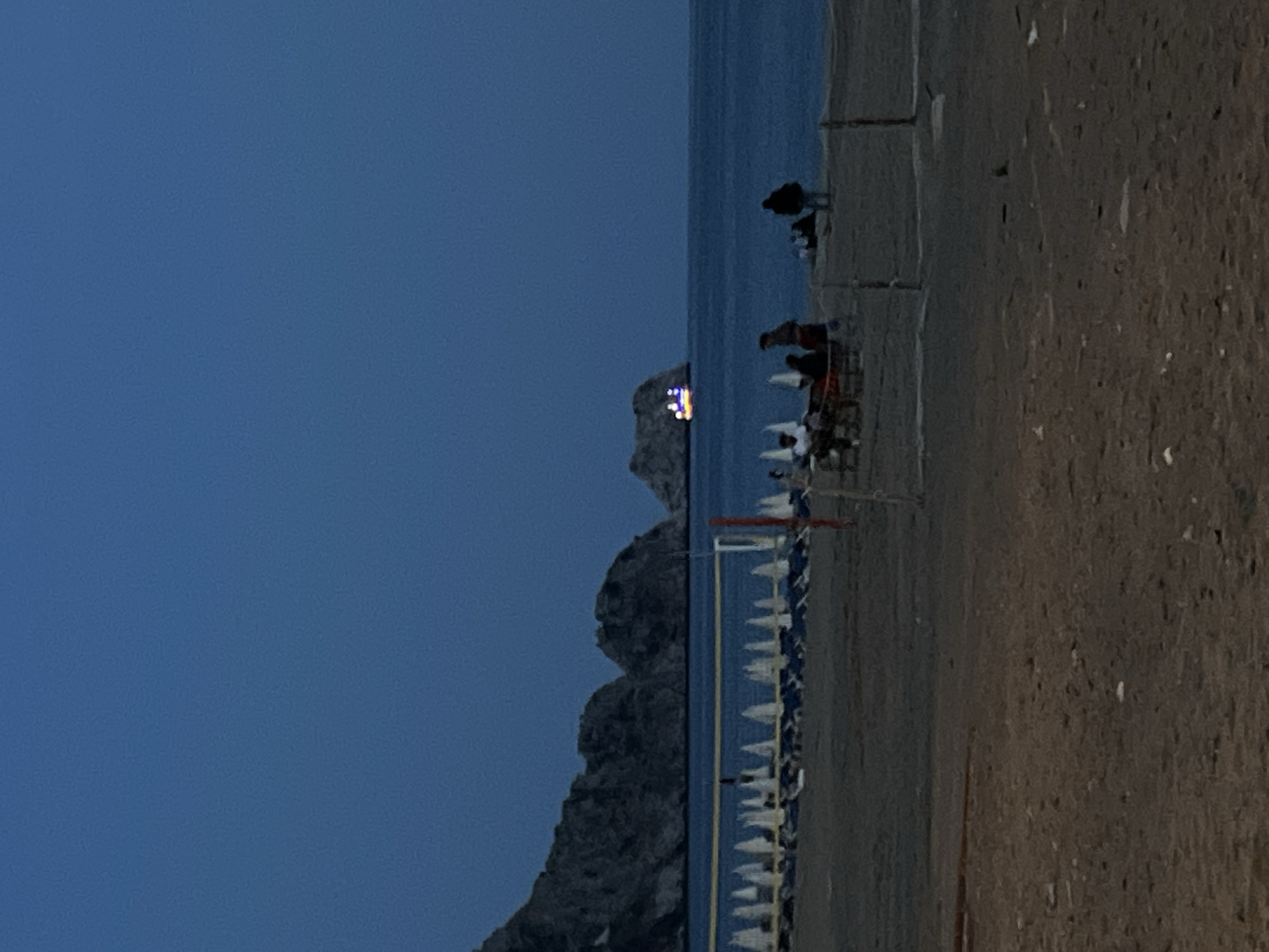 The image depicts a beach scene at night in Alanya, Turkey. A dark, rocky headland juts into the sea, partially obscuring a building or structure with illuminated lights on its peak. Along the beach, rows of white beach umbrellas stand uniformly, suggesting a resort area. Further down the beach, several people are visible, some standing near the water and others seated. The environment is defined by a dark sky, a calm sea, and a sandy beach.