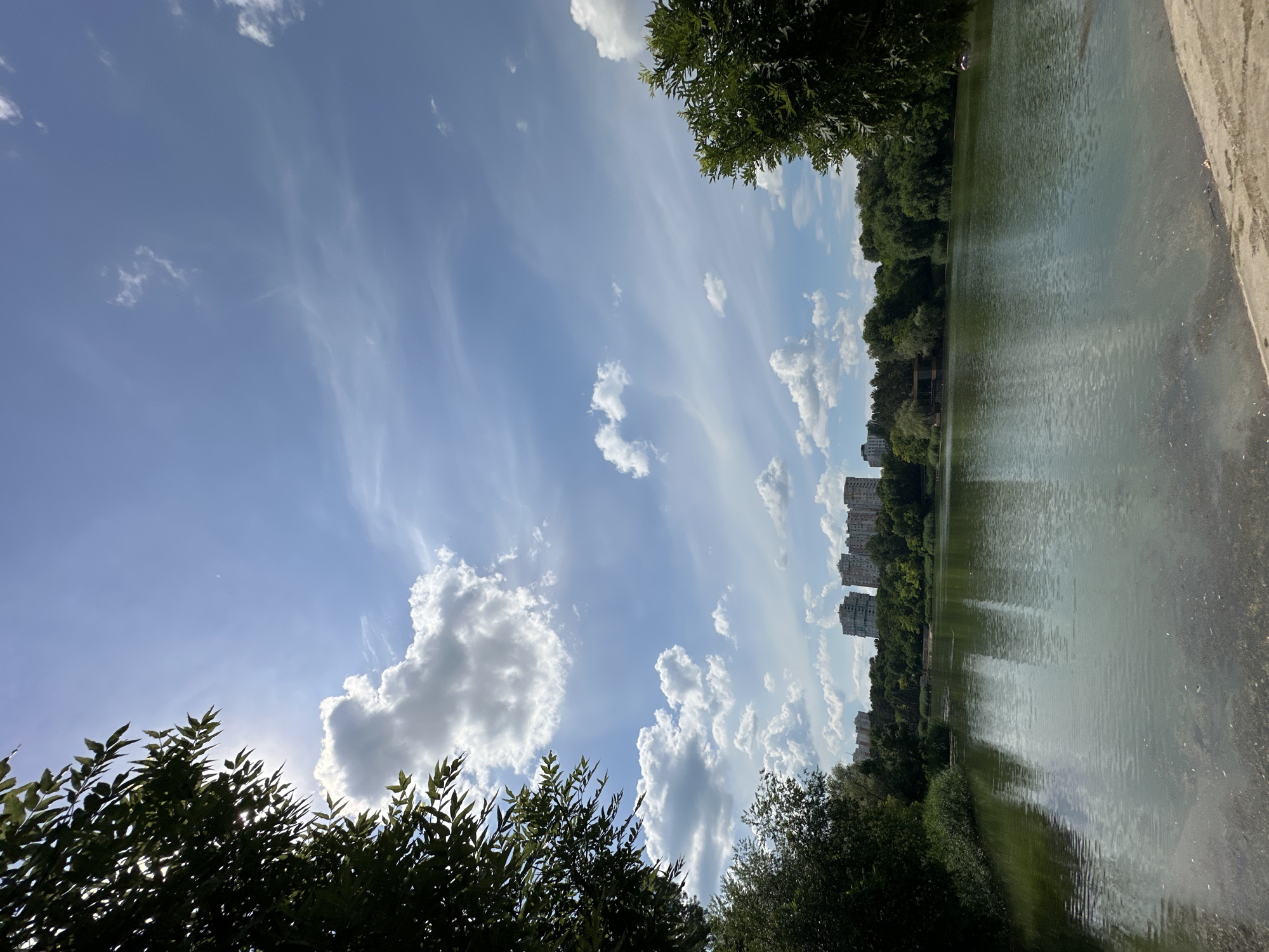 The image depicts a tranquil lakeside scene under a bright, partially cloudy sky. The perspective is from ground level, looking upwards and across a calm body of water. Dense green foliage, likely trees, frames the left and right edges of the frame, with some branches extending into the foreground. In the distance, across the lake, a line of trees forms the bank, behind which stand several multi-story residential buildings. The water itself is a muted green, reflecting the sky and the surrounding greenery. The sky is a vibrant blue, adorned with a mix of fluffy white cumulus clouds and wispy cirrus clouds, suggesting fair weather. The sun is partially visible, casting a soft glow through the clouds on the left side of the frame. The overall atmosphere is peaceful and serene, evoking a sense of calm. The presence of the buildings suggests an urban or suburban setting, likely a park or recreational area within Chișinău, Moldova. There is no visible text in the image.