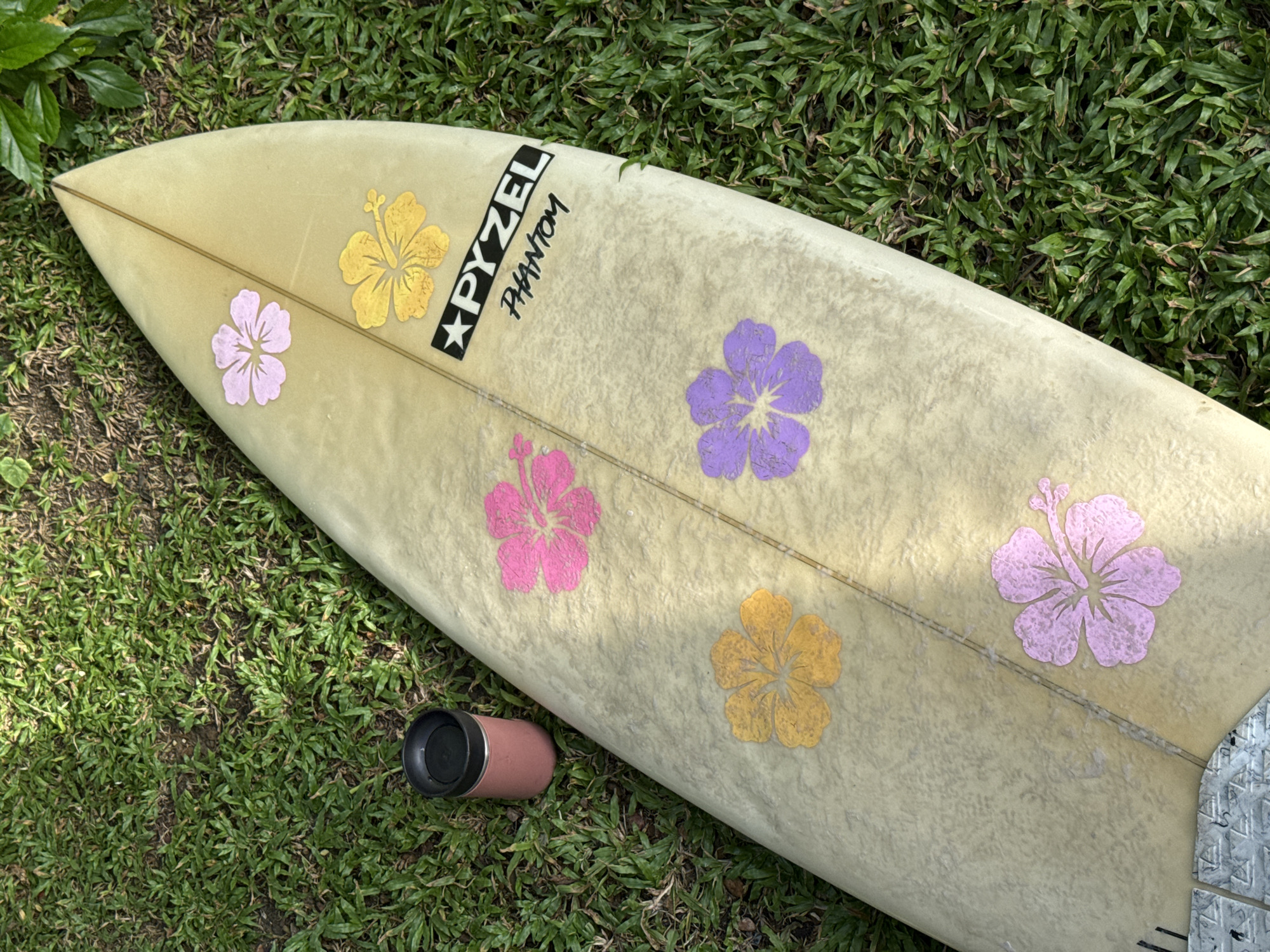 An overhead shot captures a surfboard lying on lush green grass. The surfboard is light yellow with a faint textured pattern and features five hibiscus flower stickers in pink, yellow, and purple. The brand name "PYZEL" is emblazoned in black and white on the top right of the board, with "PHANTOM" written beneath it in a script font. To the left of the surfboard, a pink insulated tumbler with a black lid sits on the grass. The background consists of more dense green grass and hints of leafy foliage, suggesting an outdoor setting in a tropical location, possibly Goviyapana, Sri Lanka.