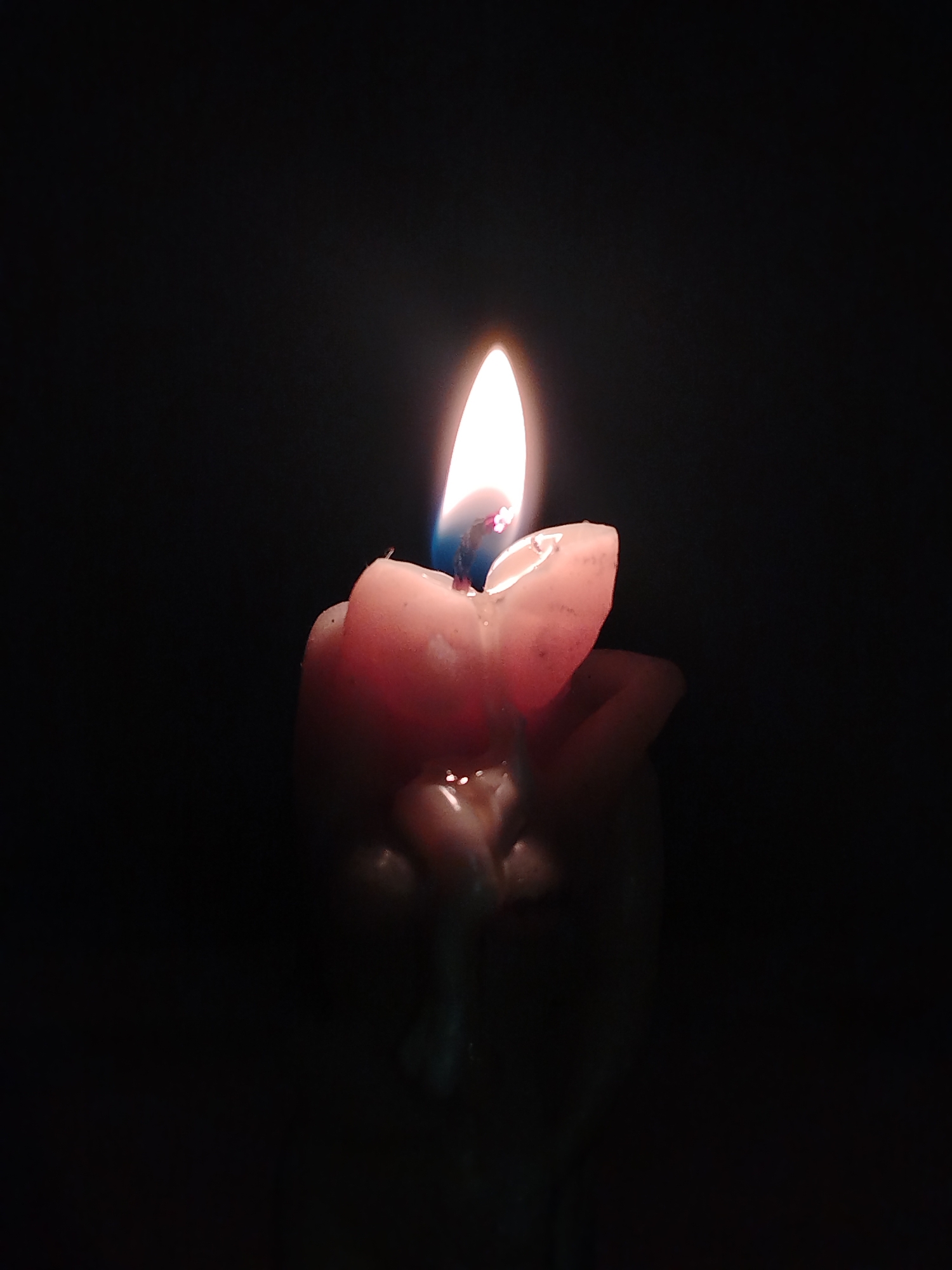 A close-up, low-lit photograph shows a burning candle. The candle itself is pinkish in color and appears to be partially melted. The flame is the primary source of light in the image, illuminating the top of the candle and casting a slight glow on the surrounding darkness. No other objects or people are visible in the frame. The location and date the image was captured cannot be identified.