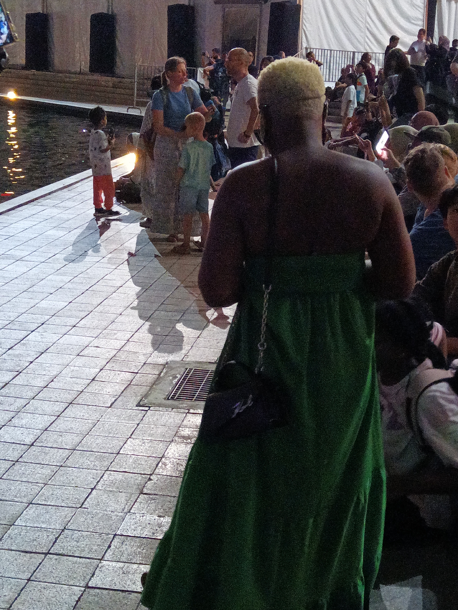 The image shows a group of people at night, possibly at an outdoor event. In the foreground, a person wearing a green dress is partially visible from behind, with their back to the camera. They are holding a dark bag with a chain strap. To the left, a young boy in an orange skirt and white shirt stands near a body of water. Another child in a light blue shirt and shorts is standing near a woman in a blue top and patterned skirt. The ground is paved with square tiles. The background is blurred, showing a crowd of people and some structures. The overall scene suggests a public gathering in a city setting. Without further context or identifiable landmarks, it is impossible to determine the specific location within Kuala Lumpur, Malaysia.